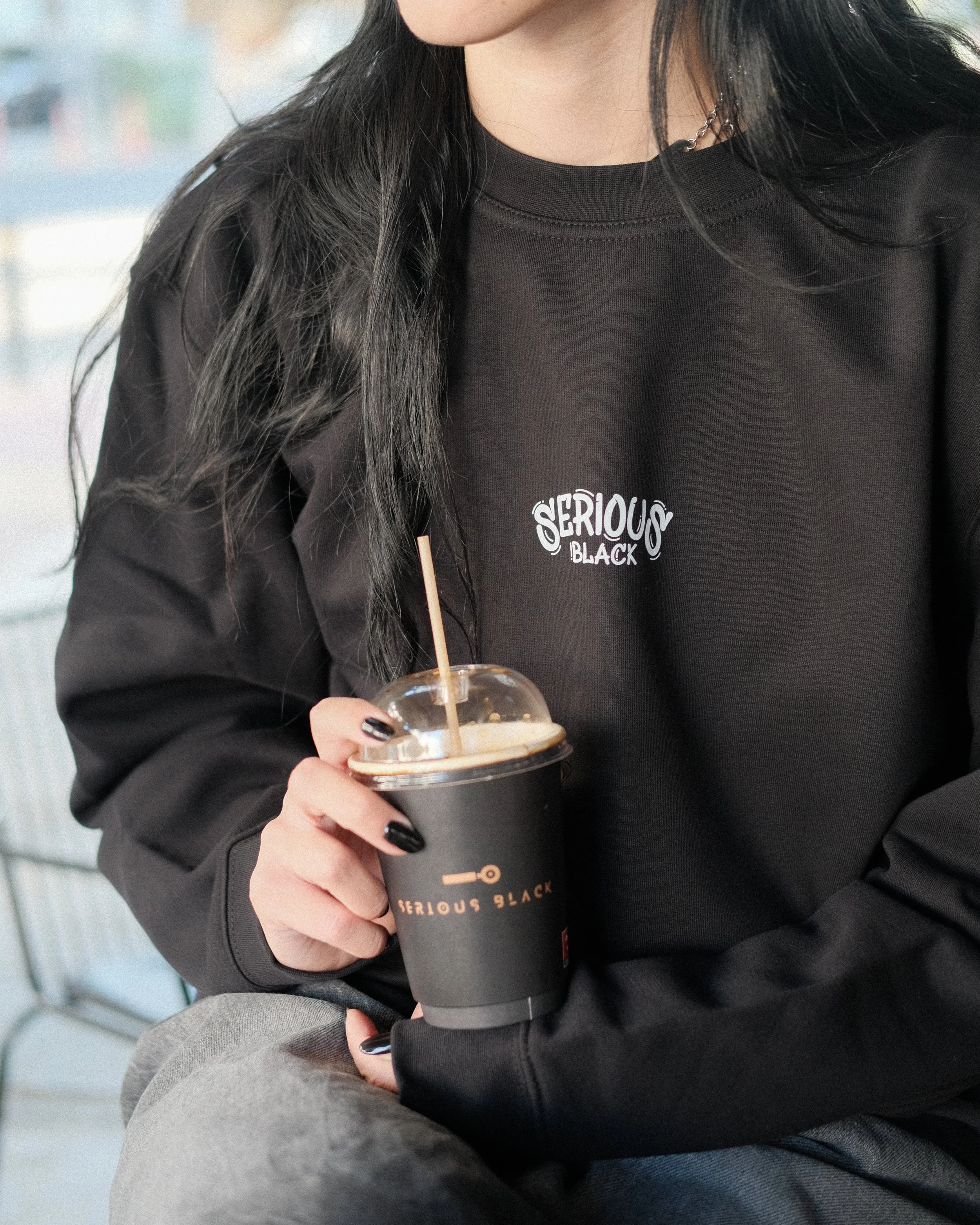 A woman with long black hair, wearing a black sweatshirt with the words 'Serious Black' on it, holding a black disposable coffee cup with a clear plastic lid and straw, sitting outdoors.