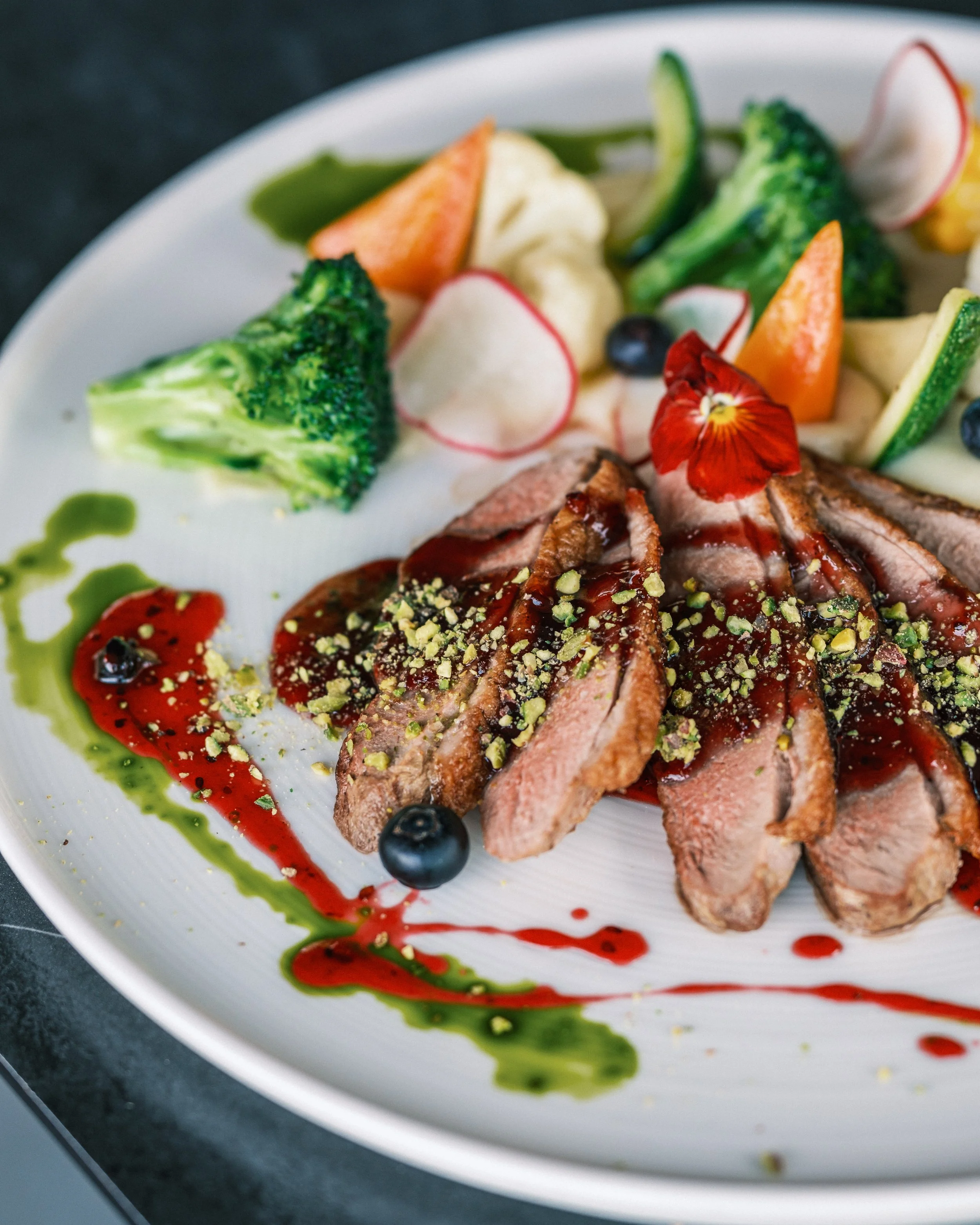 Sliced steak with a berry sauce and crushed pistachios, garnished with edible flowers, served with broccoli, cucumber, radish, carrots, and blueberries, drizzled with green and red sauces on a white plate.