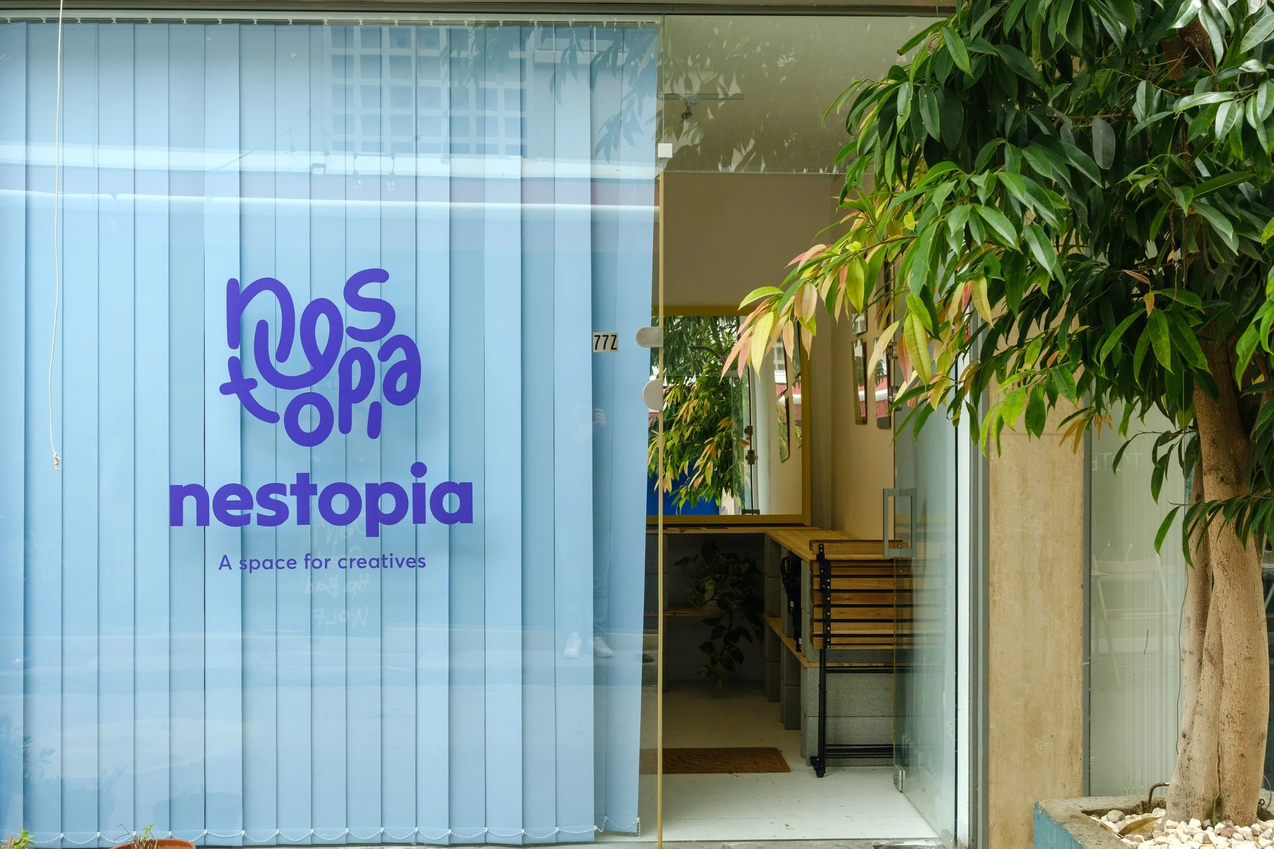 Storefront with a blue glass door that has purple text reading "nestopia" and a subtitle "A space for creatives." There is a shop interior with wooden shelves visible through the door, and a tree with green leaves outside on the right.