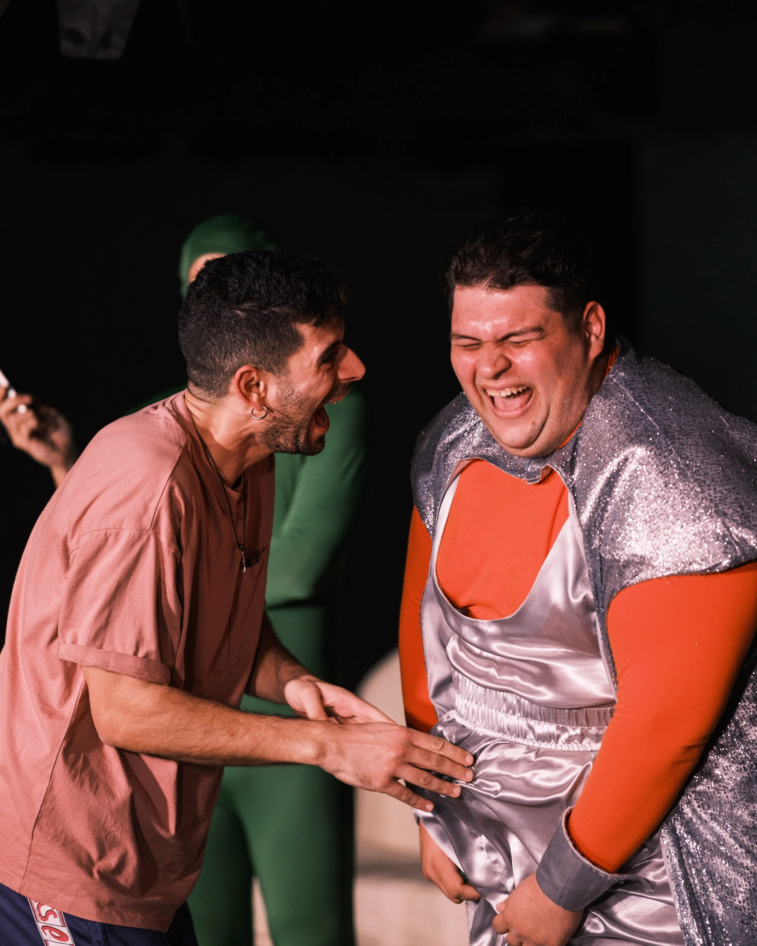 Two men happily laughing and sharing a moment, one dressed as a boxer or wrestler in a shiny silver and orange outfit, and the other in a mauve shirt, both with dark hair, against a dark background.