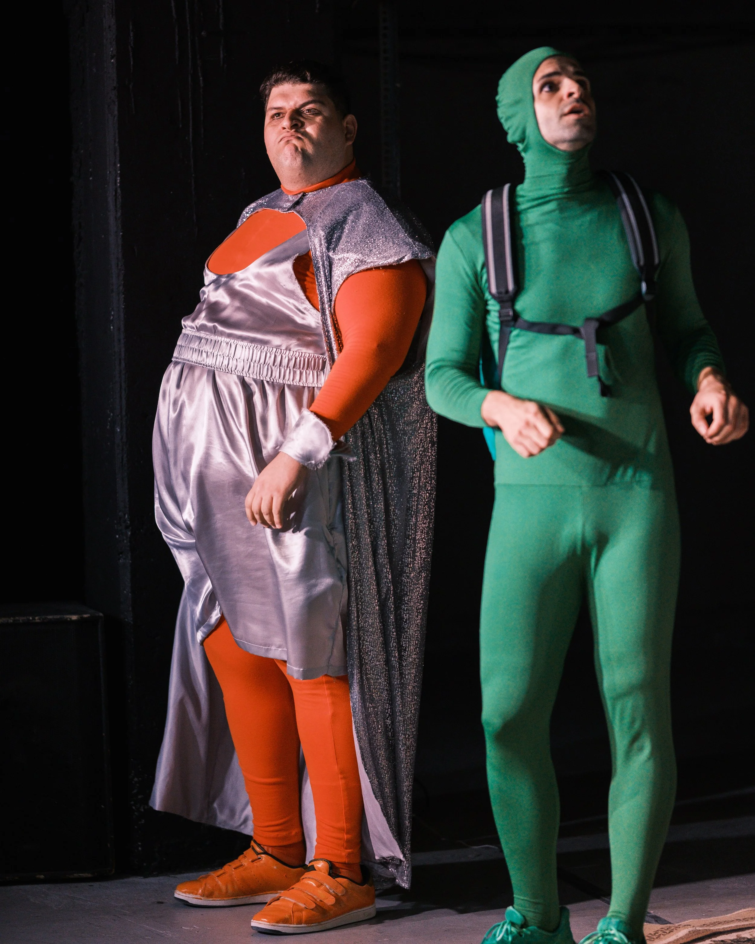 Two men standing side by side in costumes, one in an orange, silver, and pink space-themed outfit with matching sneakers, and the other in an entirely green outfit with a head covering and backpack, in a dark setting.