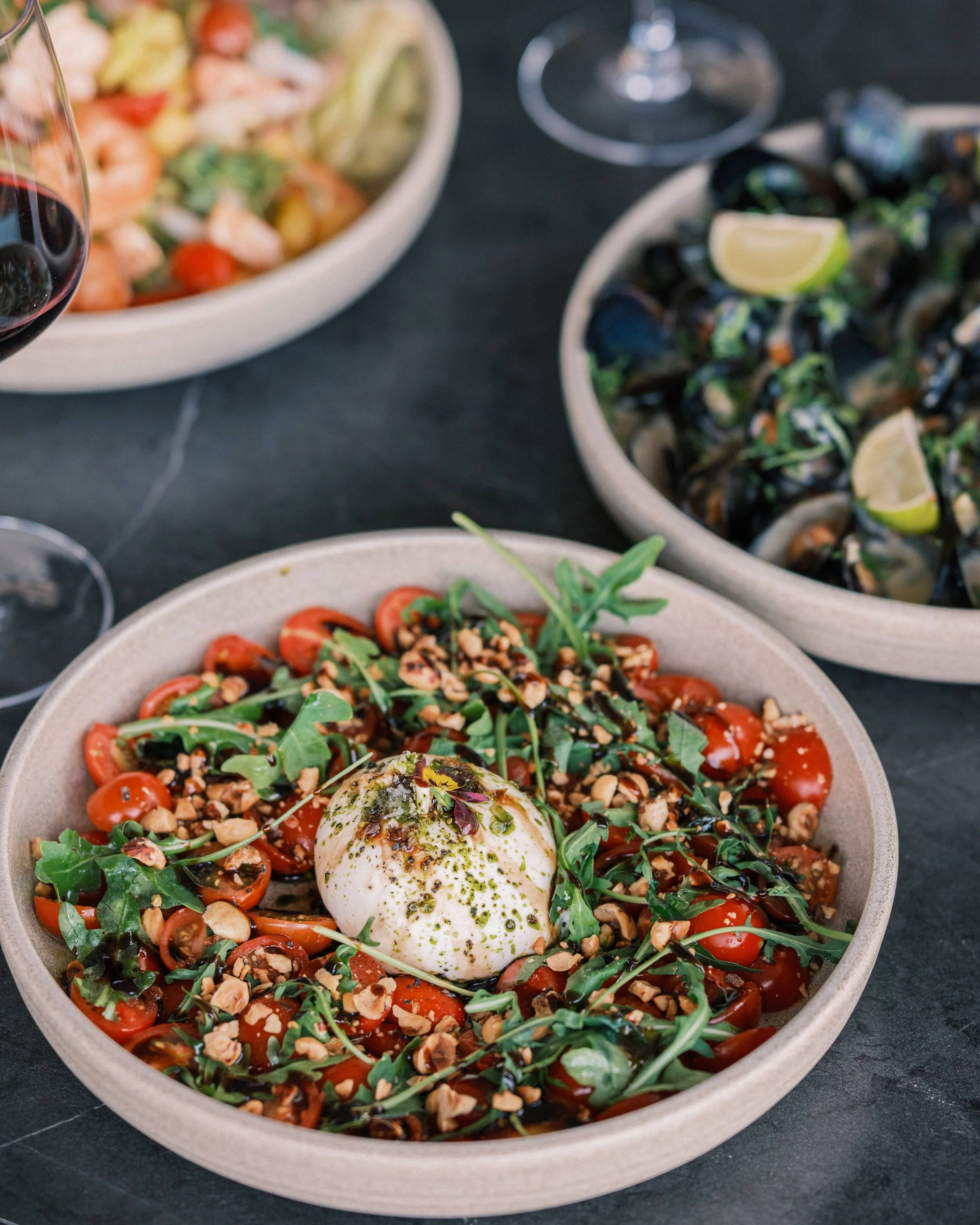 A bowl of cherry tomato salad garnished with chopped nuts, arugula, and a scoop of burrata cheese, drizzled with balsamic and herbs, on a black table with other bowls of salad and a glass of red wine in the background.