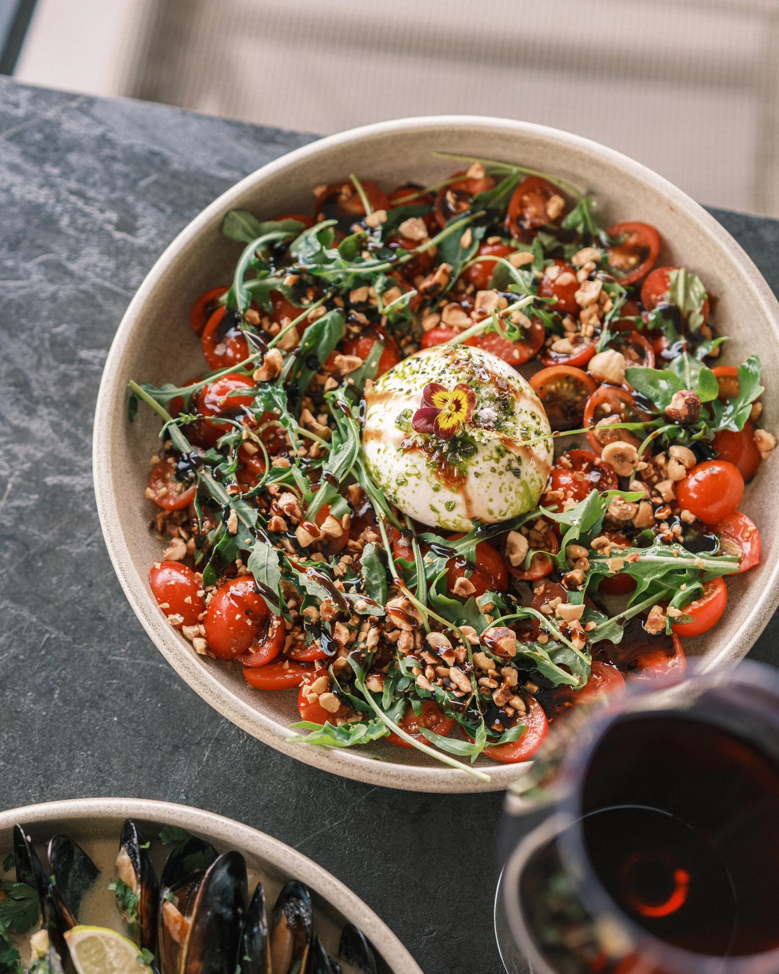A close-up of a bowl filled with cherry tomatoes, arugula, chopped nuts, balsamic glaze, and a poached egg topped with edible flowers, on a dark countertop.