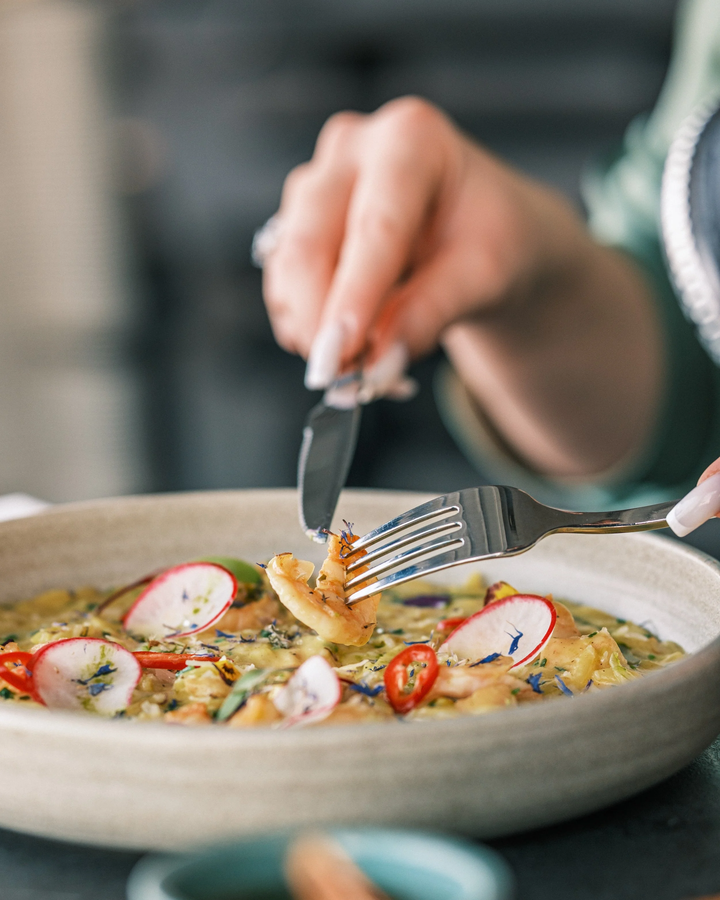 Close-up of a fork picking up a piece of pasta with colorful edible flowers and sliced vegetables on a pasta dish on a white plate