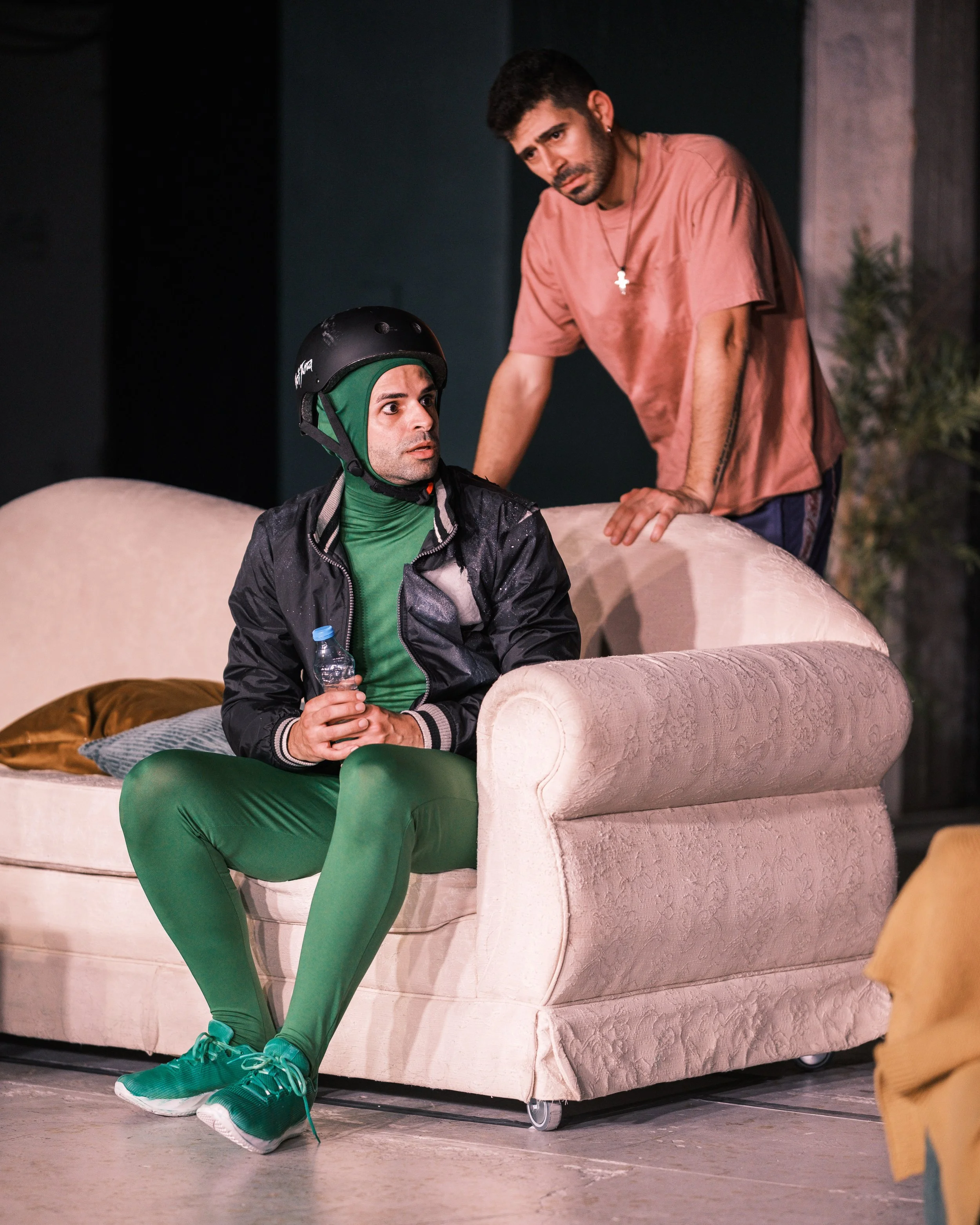 Two men on a stage. One sitting on a beige vintage sofa, holding a water bottle, wearing green leggings, sneakers, a black jacket, and a helmet. The other man stands behind the sofa, leaning on it, wearing a pink t-shirt and necklace. The background 