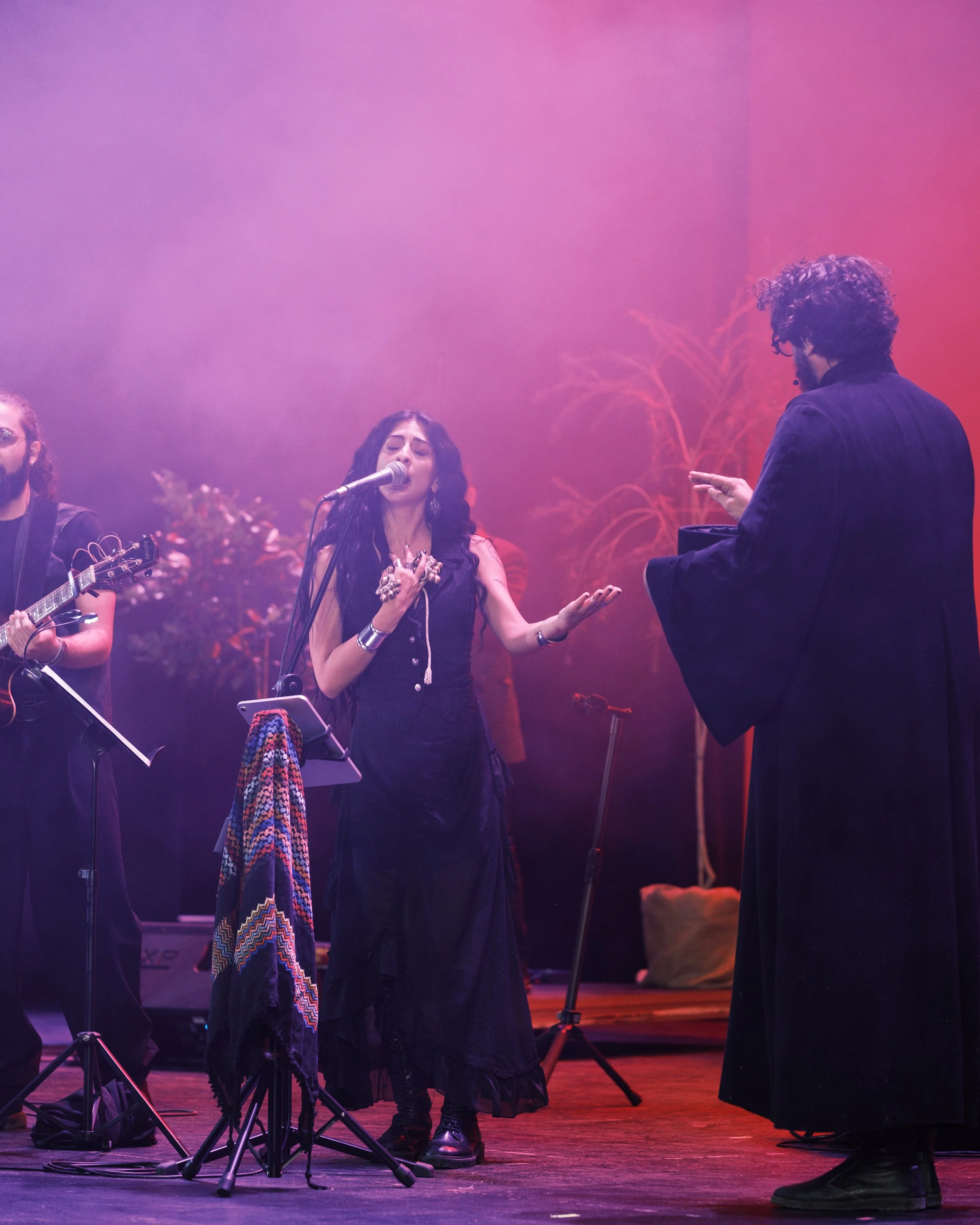 Musicians performing on stage with a woman singing into a microphone, a man playing guitar, and another man conducting, with purple and pink lighting and autumn-themed decorations in the background.