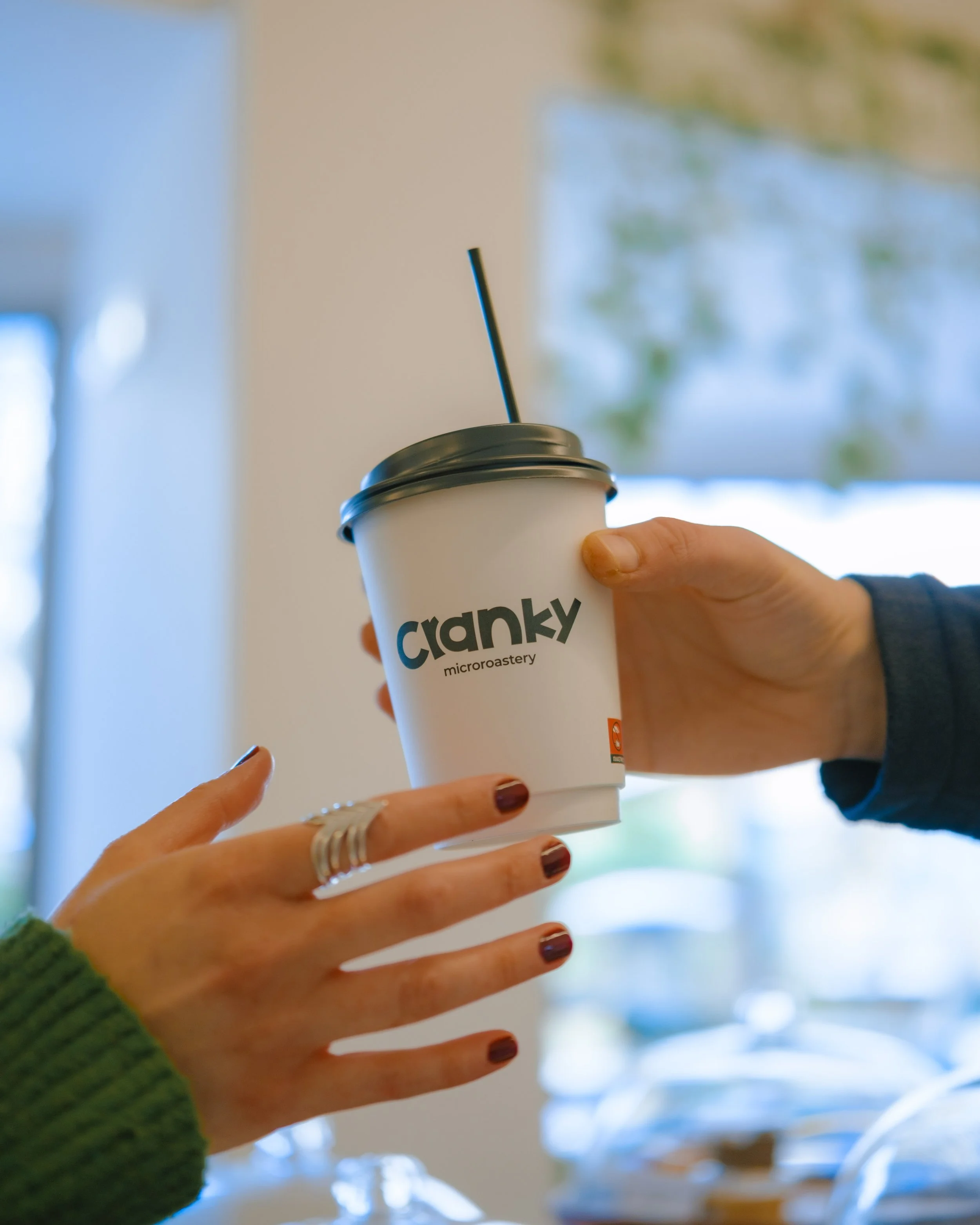 Person holding a cup with a straw, the cup has the logo 'cranky micro roastery' on it, in an indoor setting with blurred background.