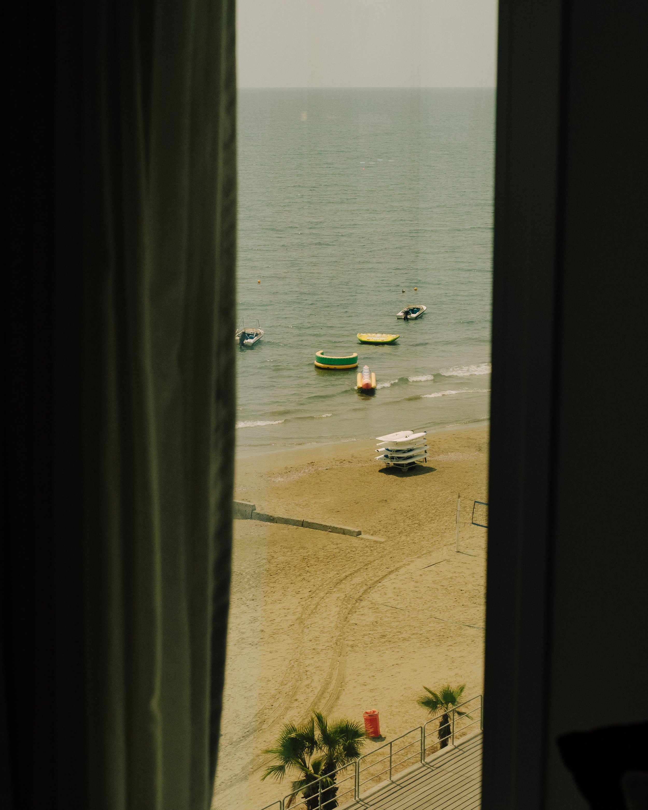 View of a beach with boats floating on the water, seen through a window with green curtains, with palm trees and a sandy shore in the foreground.