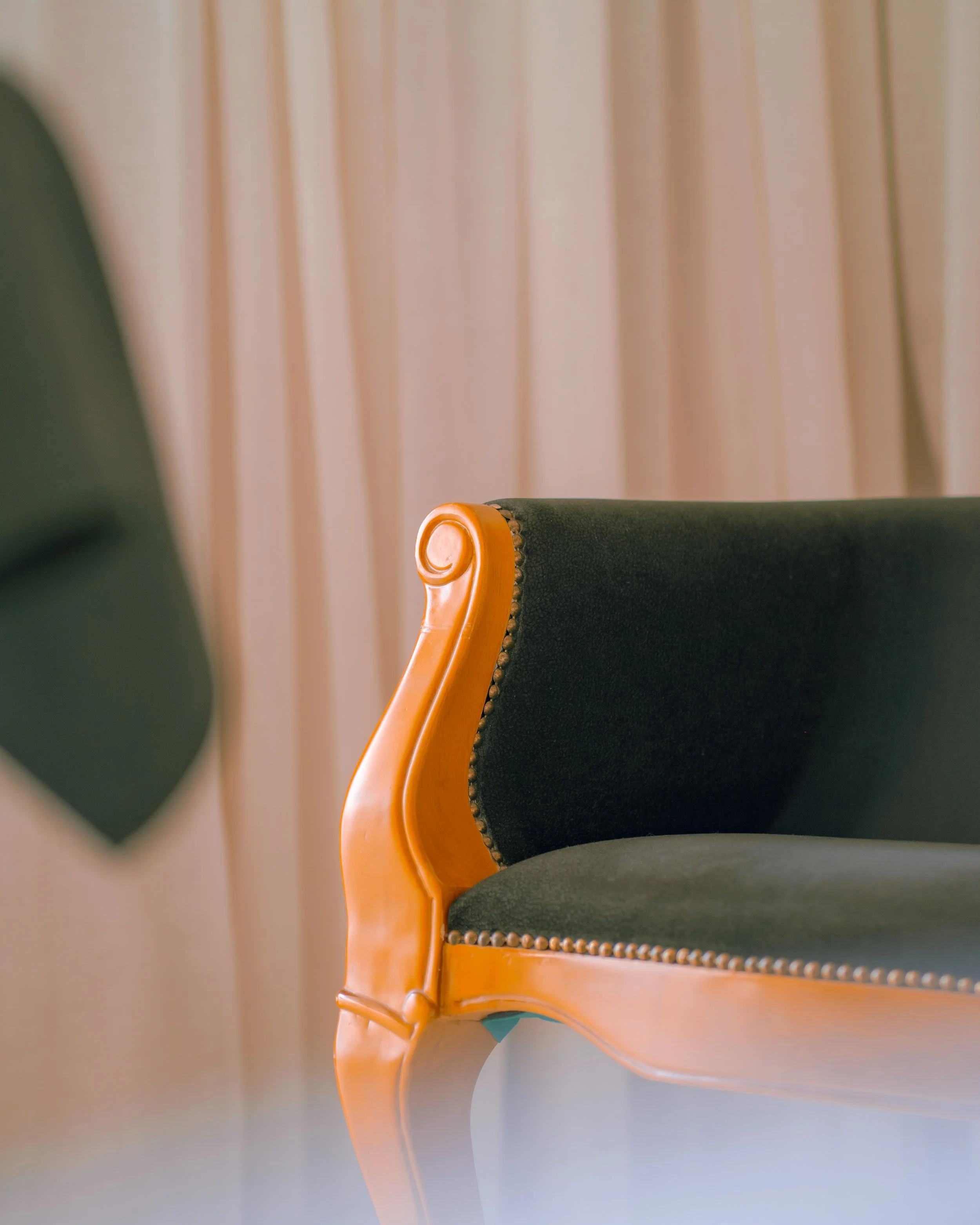 Close-up of the side of a wooden and black upholstered chair with curved armrest and decorative studs, against a background of beige curtains.