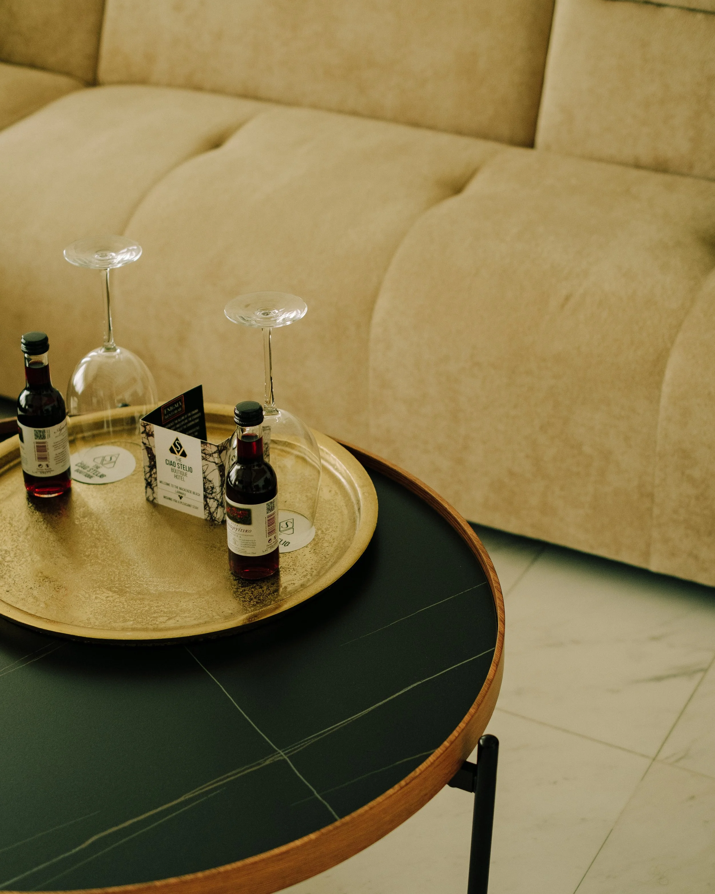 A table with a gold-colored surface holding three empty wine glasses and three small bottles. Part of a sign with hotel information is also visible on the table. In the background, a beige upholstered headboard and part of a bed are seen.