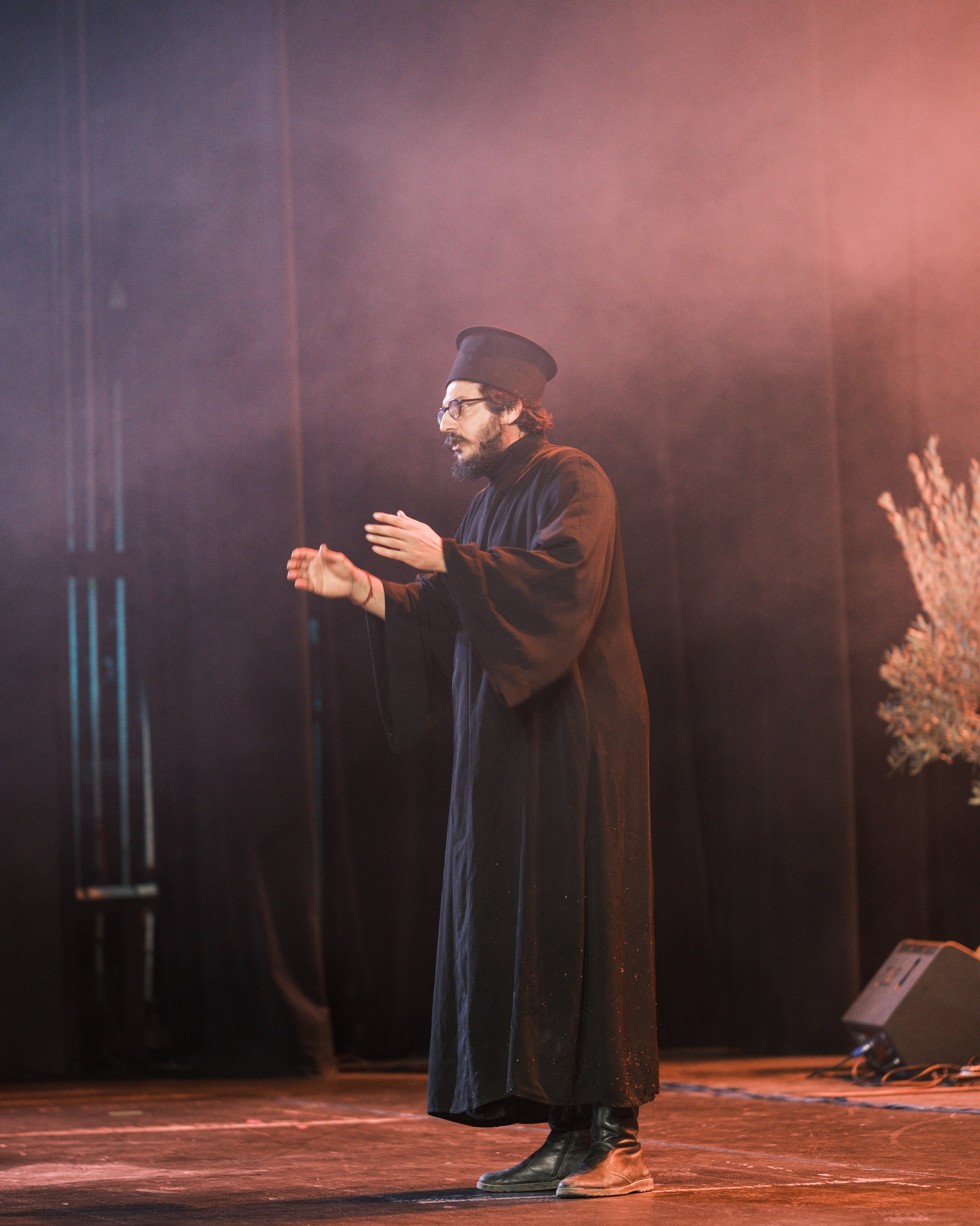 A person standing on a stage, wearing black clothing, glasses, a tall black hat, and brown boots, with a blurred background and some stage equipment visible.