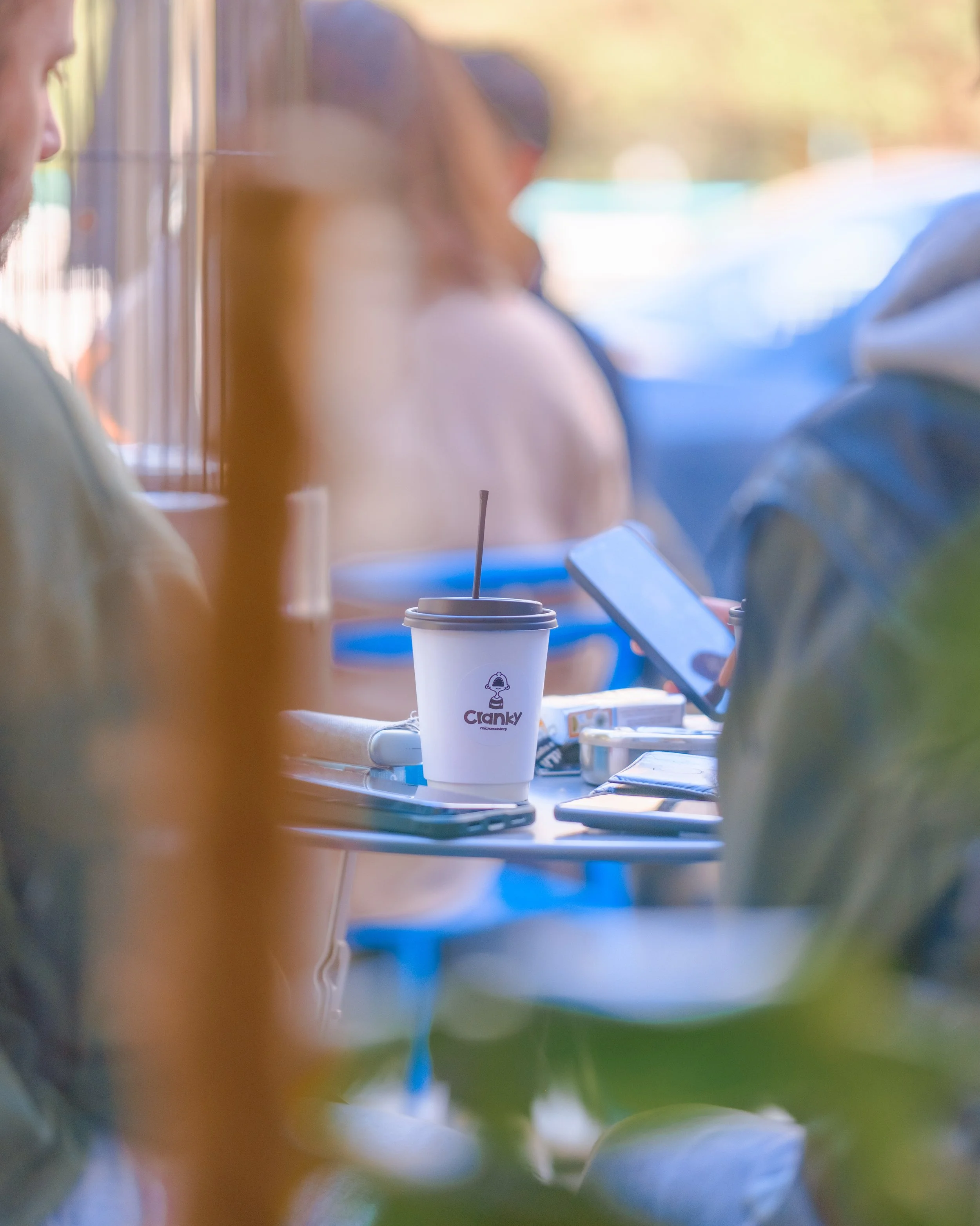 A close-up of a white to-go coffee cup with the logo 'Cranky' on it, placed on a table with two smartphones and a snack box, with blurred people in the background.