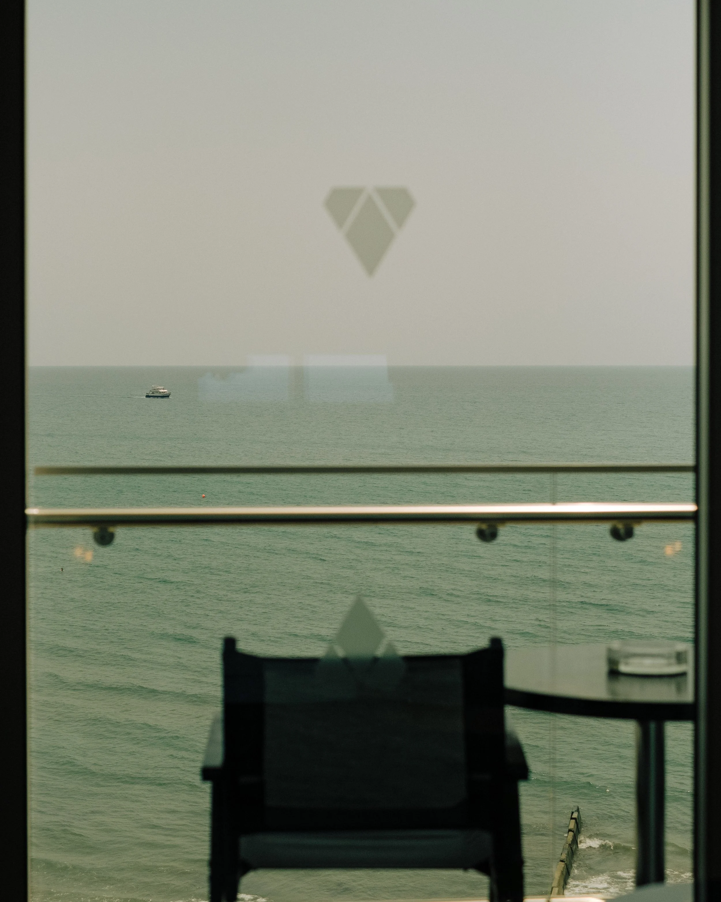 Photo of a boat on the ocean seen through a window with reflections of the room's interior, including chairs and a railing.