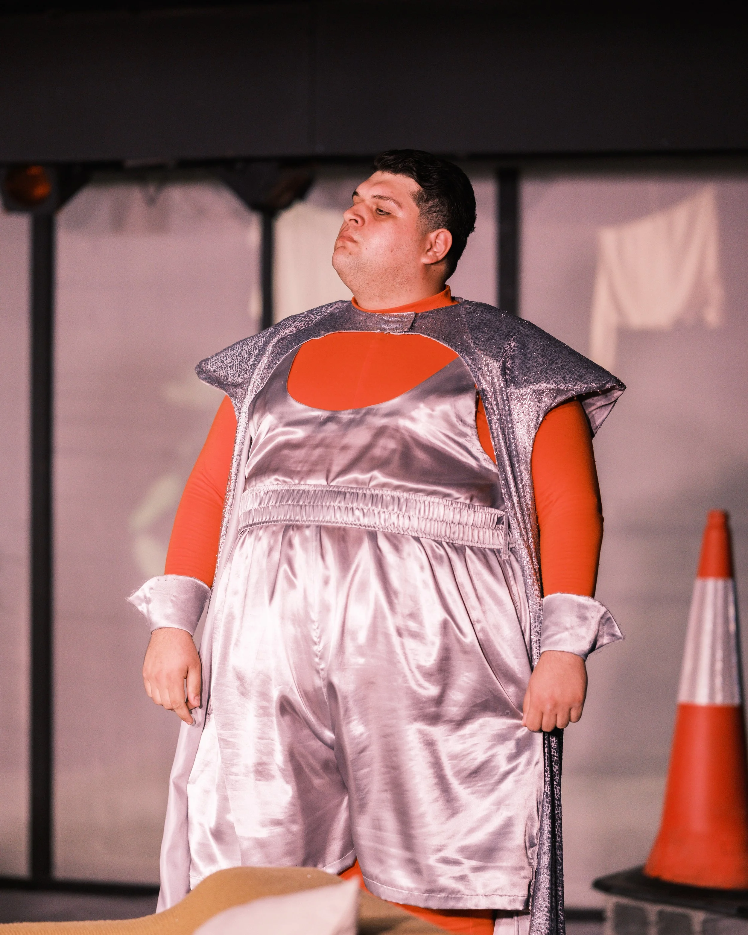 A man in superhero costume on a set, standing with eyes closed, arms relaxed, with orange traffic cones and equipment in background.