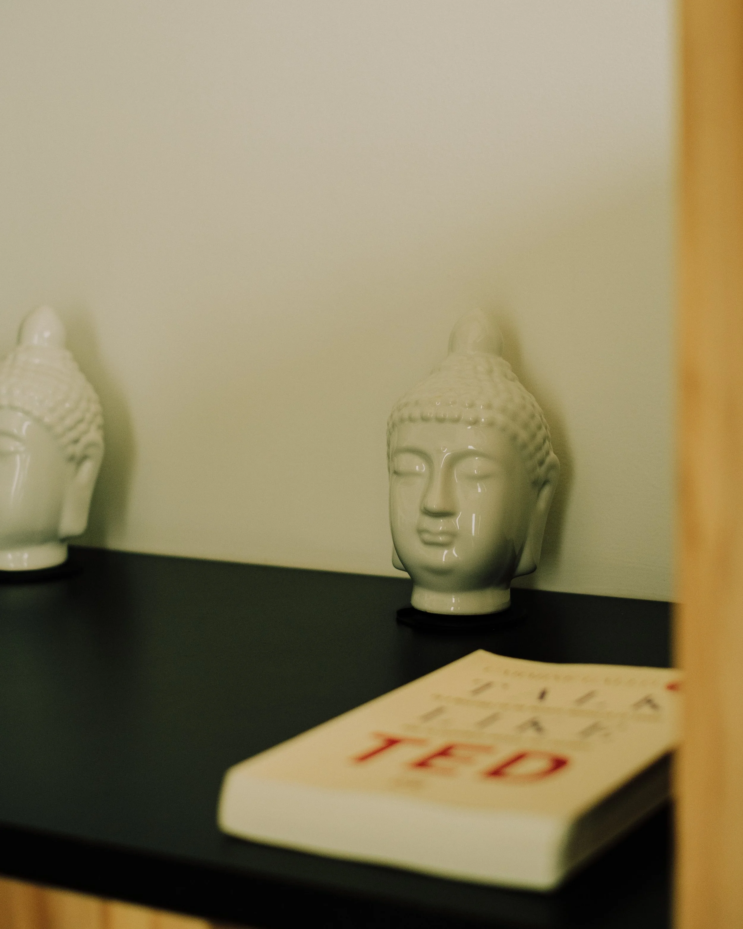 Close-up of two ceramic Buddha head statues on a dark surface against a beige wall, with a blurred card with red text in the foreground.