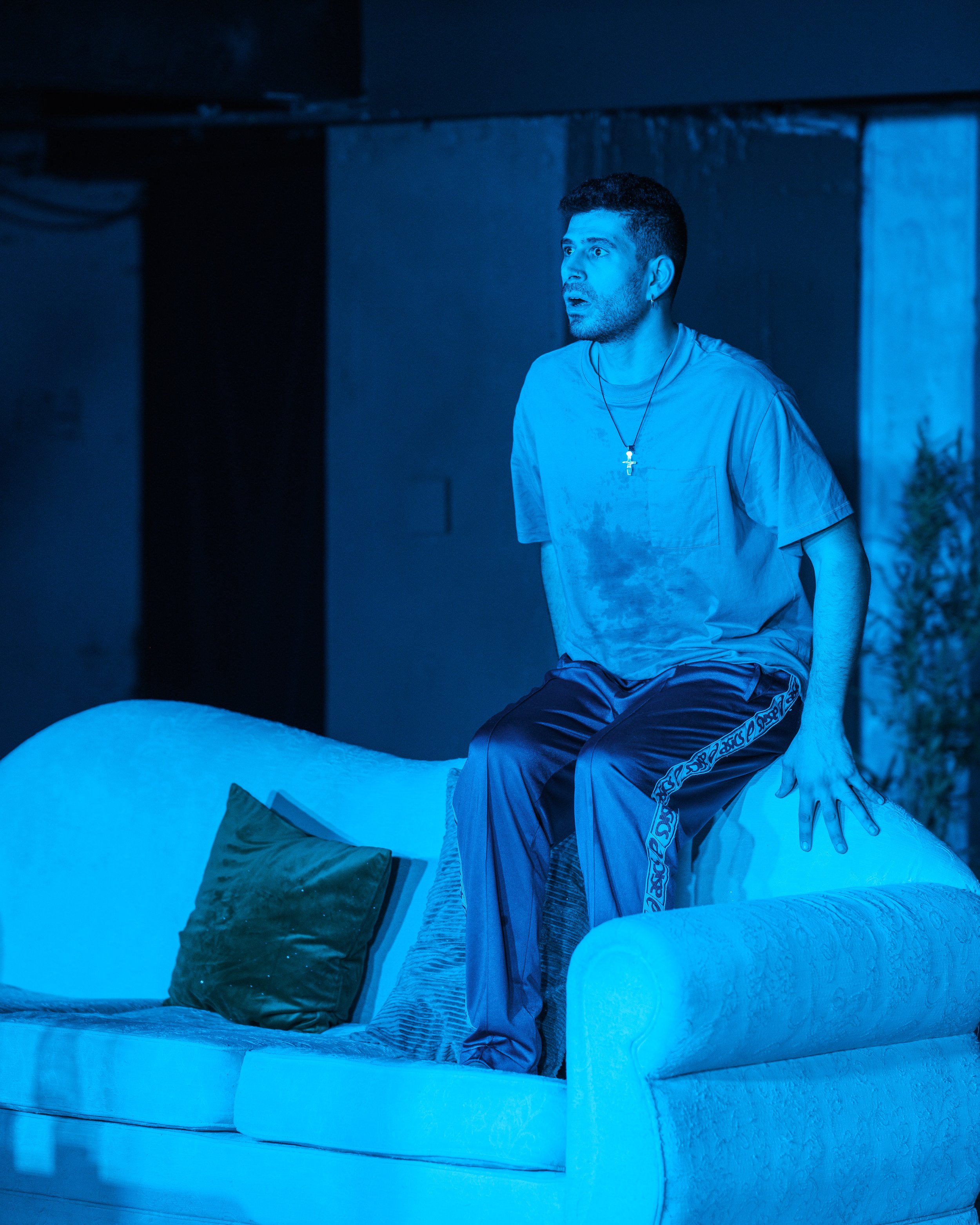A man with short dark hair and a beard, wearing a gray t-shirt and a cross necklace, sitting on an icy surface in a room illuminated with blue light, with a surprised or shocked expression on his face.