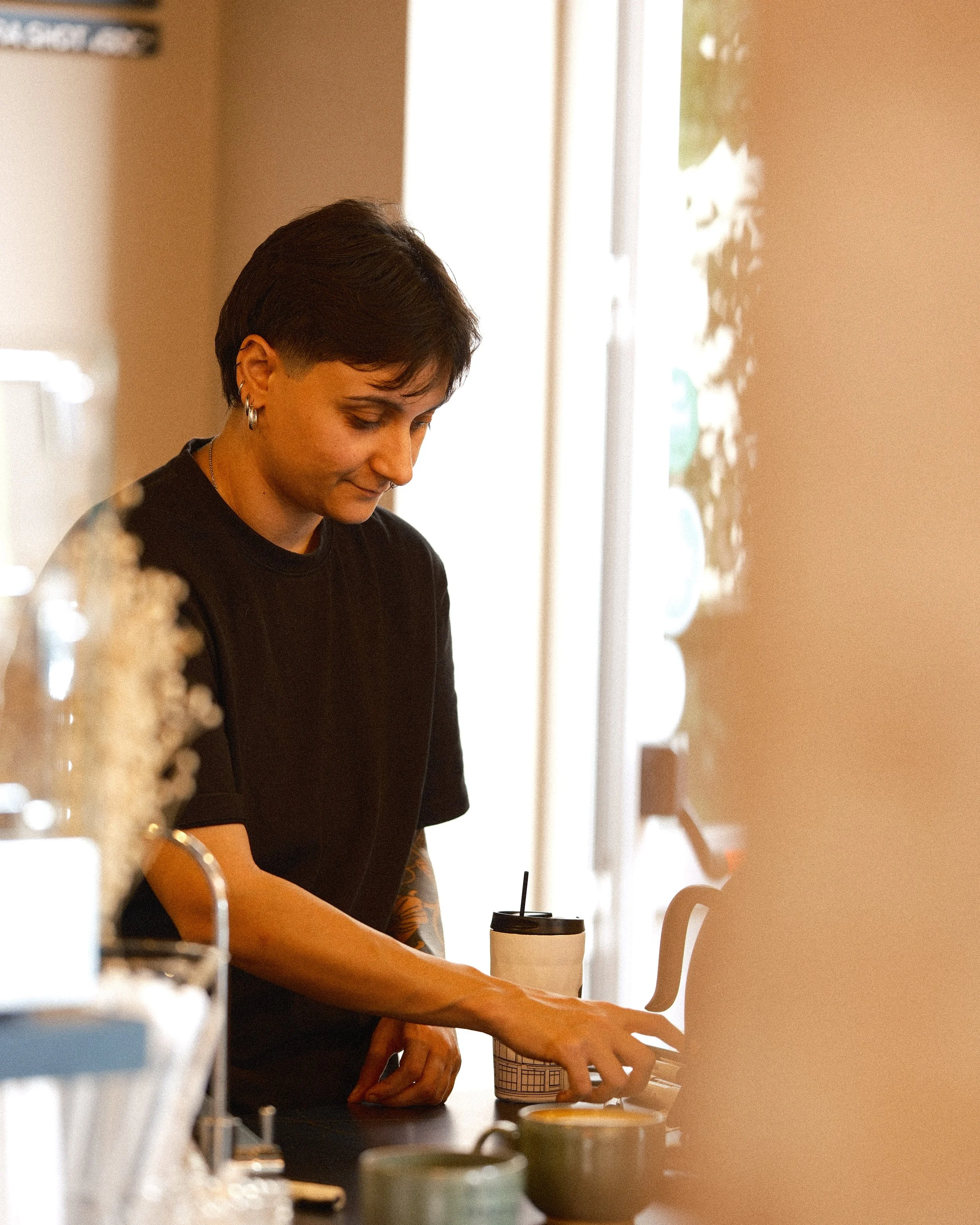 A woman is preparing coffee or tea at a counter, holding a paper cup with a lid. She has short dark hair, tattoos on her arm, and is wearing earrings and a dark top. The setting appears to be a cafe or kitchen with warm lighting, and there are blurre