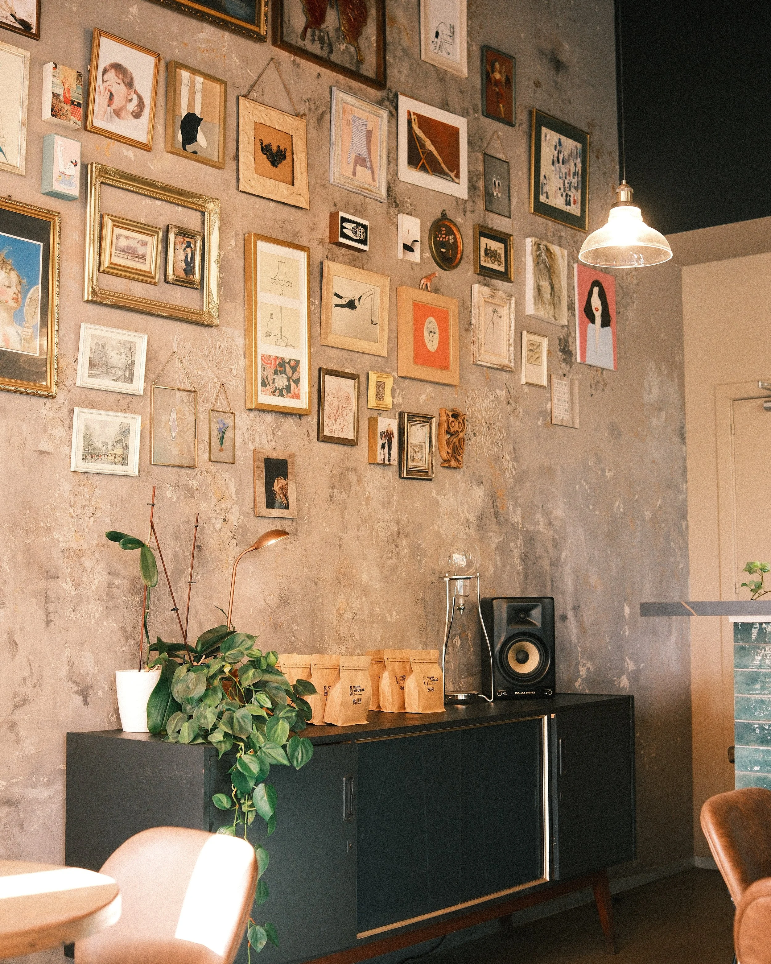 Interior of a café or studio with a textured grey wall decorated with numerous framed art prints and photographs, a black sideboard with a potted plant on the left, three brown paper coffee bags on top, and a speaker on the right, with a pendant ligh