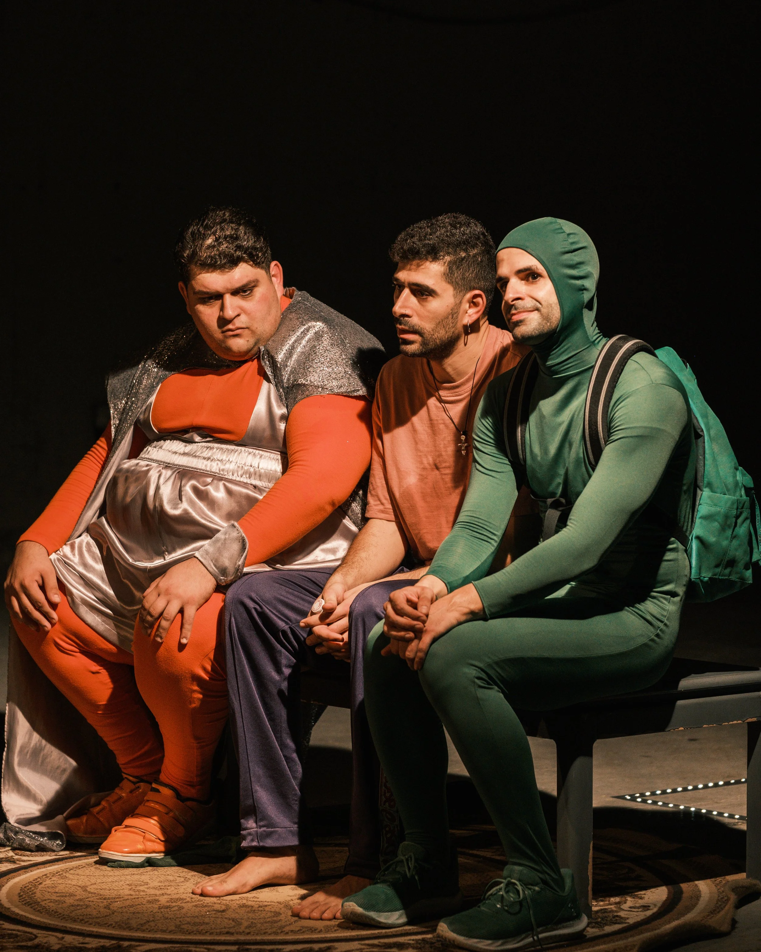 Three men sitting closely together on a stage, all wearing colorful costumes, with a dark background and a patterned rug beneath their feet.