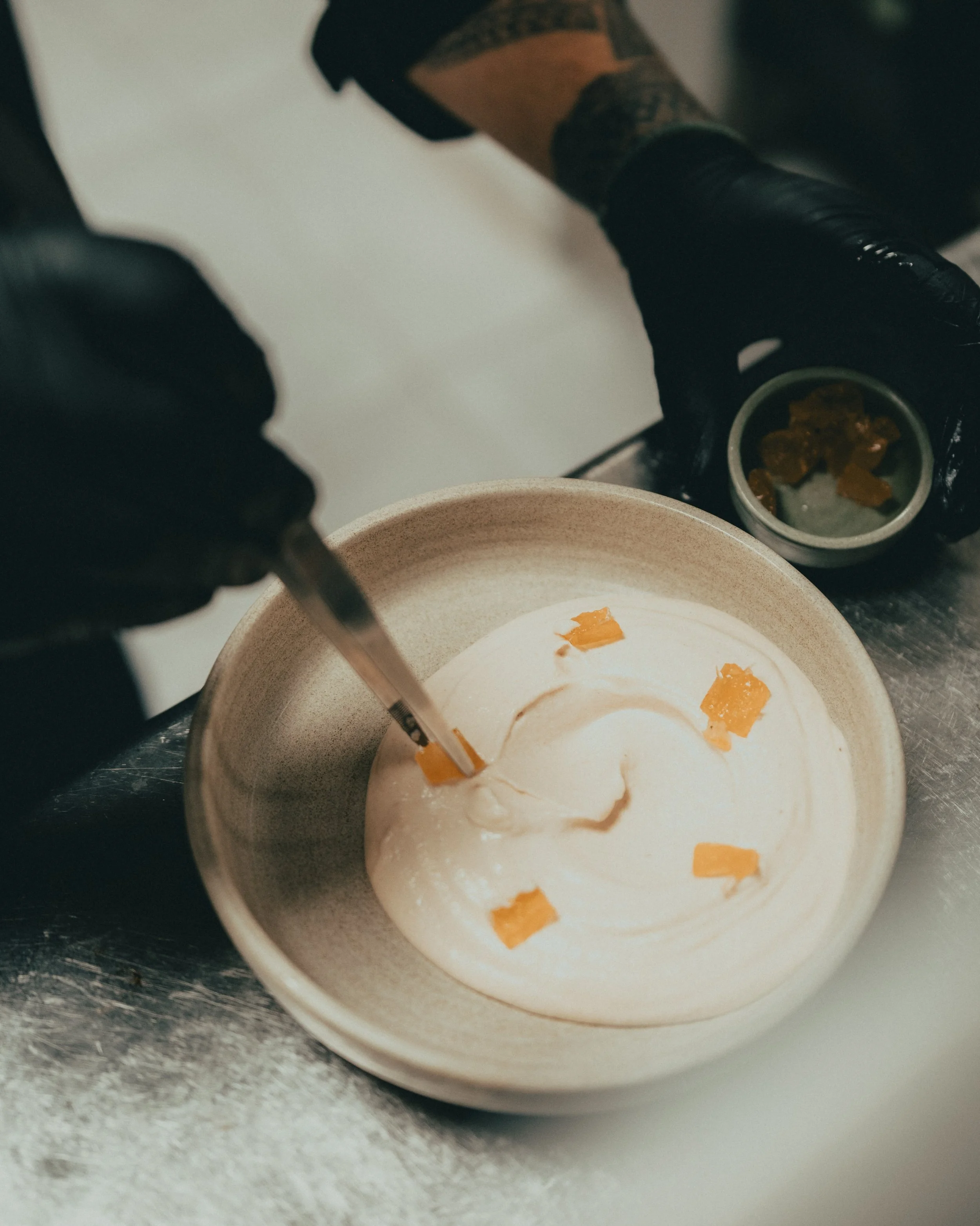Close-up of a bowl of creamy dessert garnished with orange zest, with a person wearing black gloves and lace socks in the background.