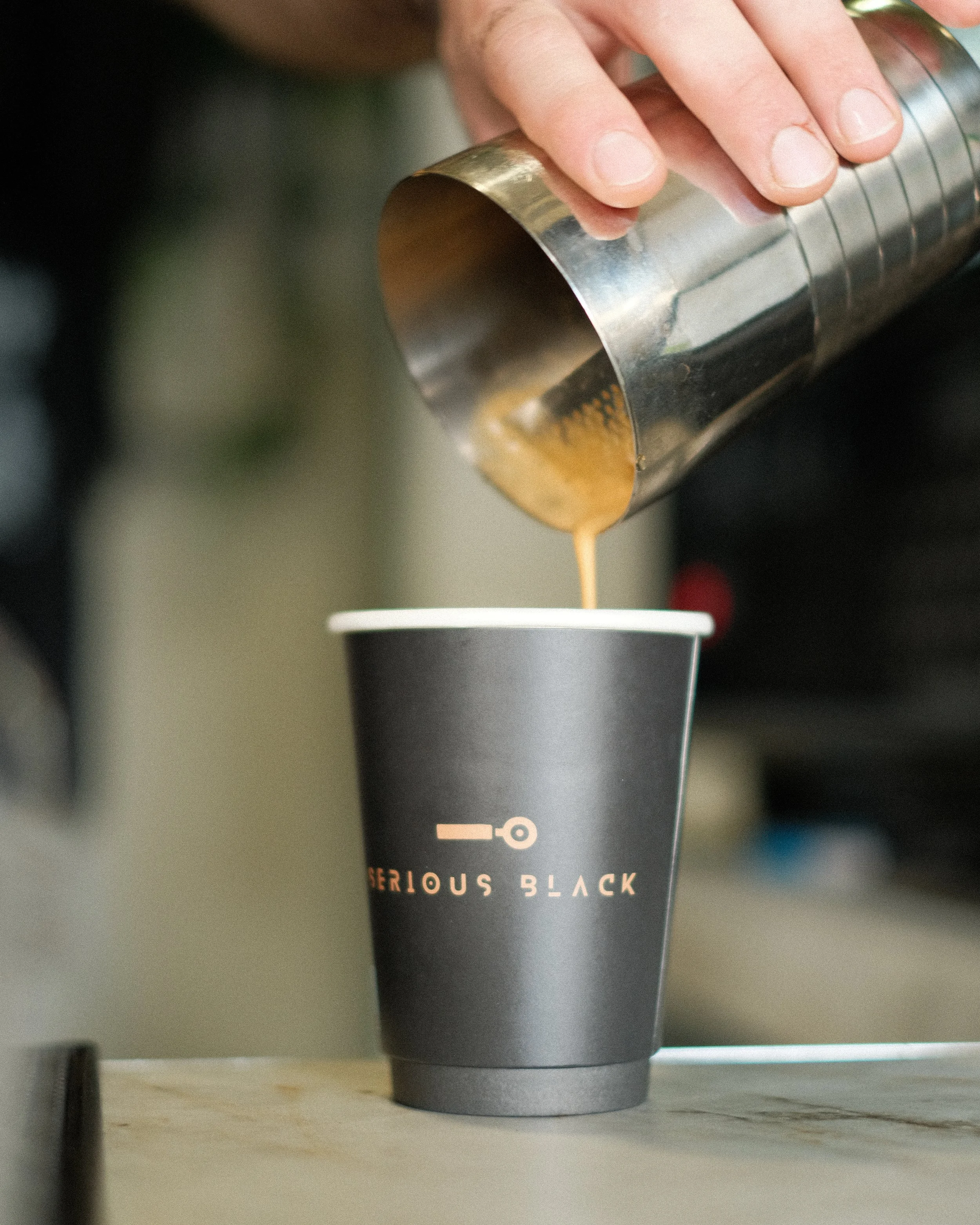 A hand holding a stainless steel shaker pouring coffee into a black cup labeled 'SERIOUS BLACK'.