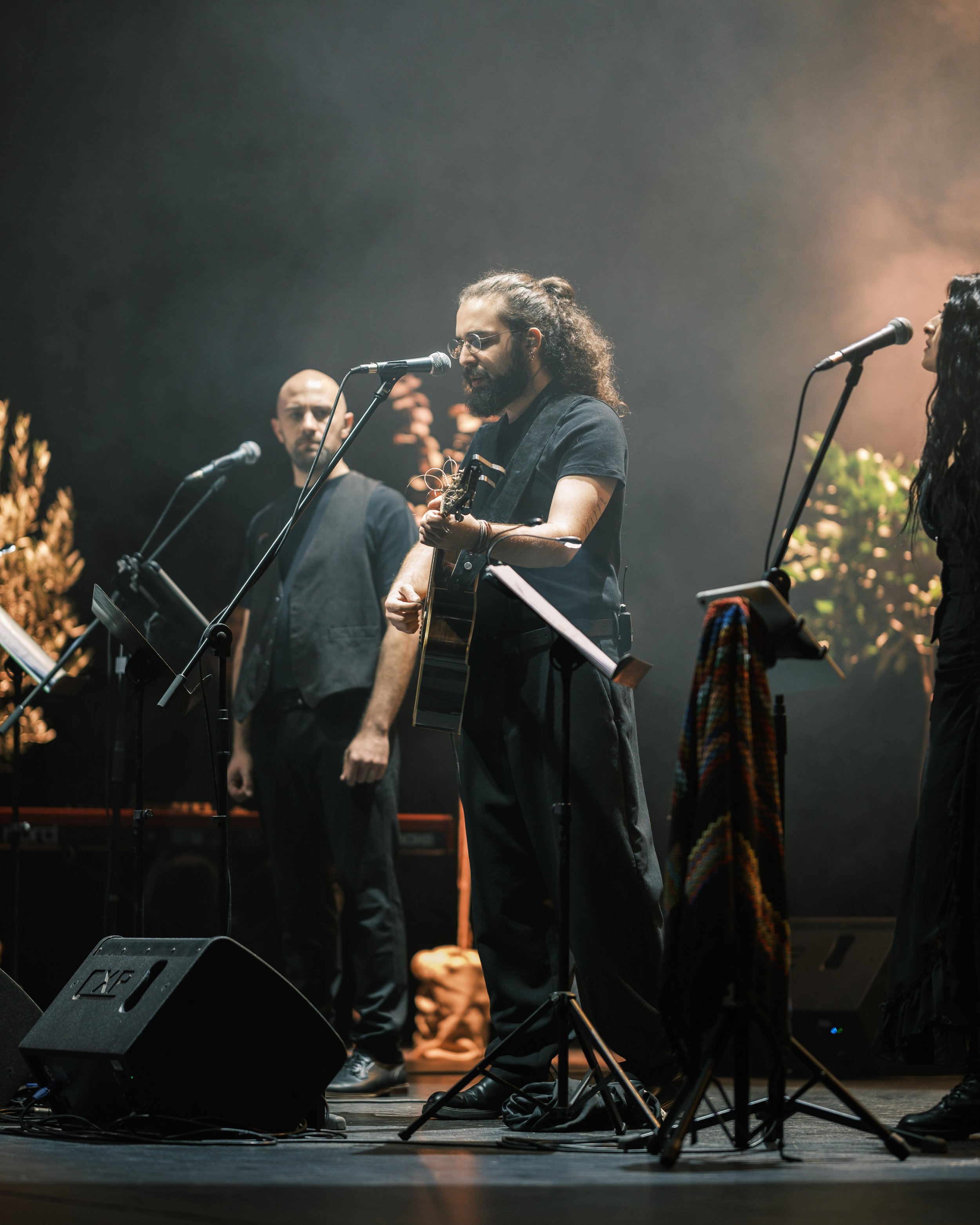 A group of musicians performing on stage, with a male singer with long curly hair and glasses singing into a microphone while playing an acoustic guitar. Other band members are visible, with musical equipment and microphones set up around them.