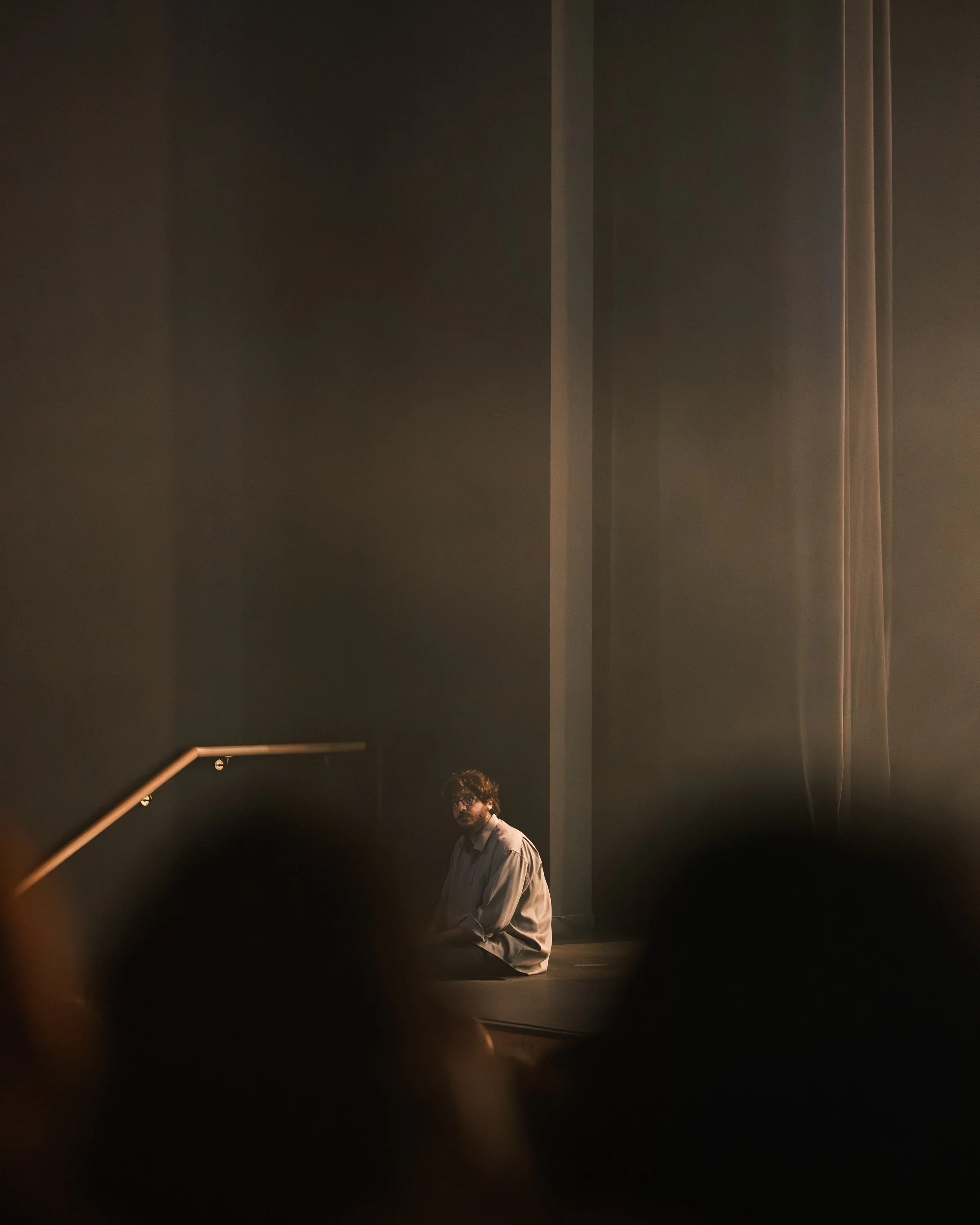 A man with glasses and curly hair is kneeling on a dark stage, wearing a white shirt, illuminated by soft lighting in a minimalist setting with dark walls and a staircase railing.