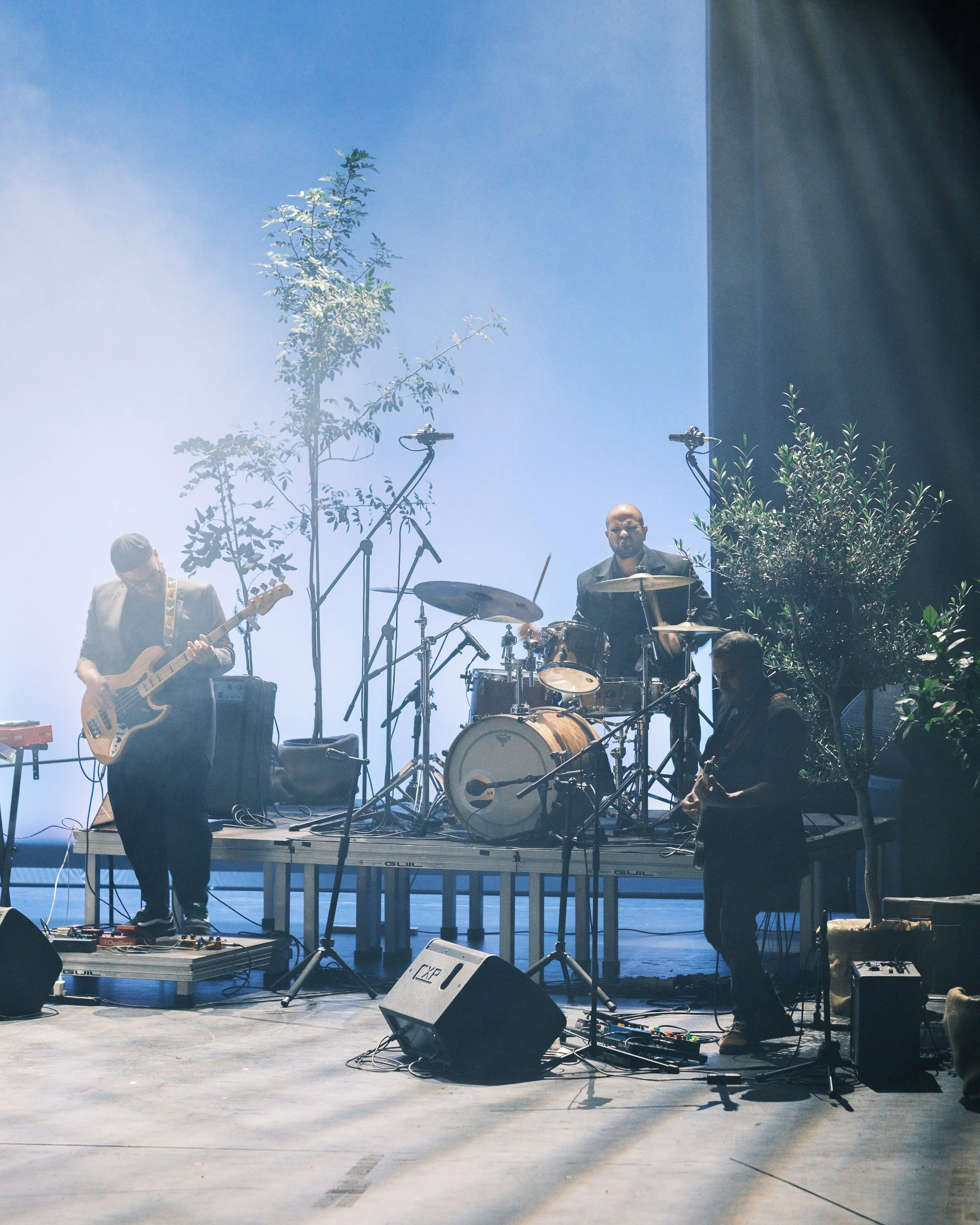 A band performing on stage with musicians playing guitar, drums, and keyboard. There are microphones and music equipment, with some plants and blue sky visible.