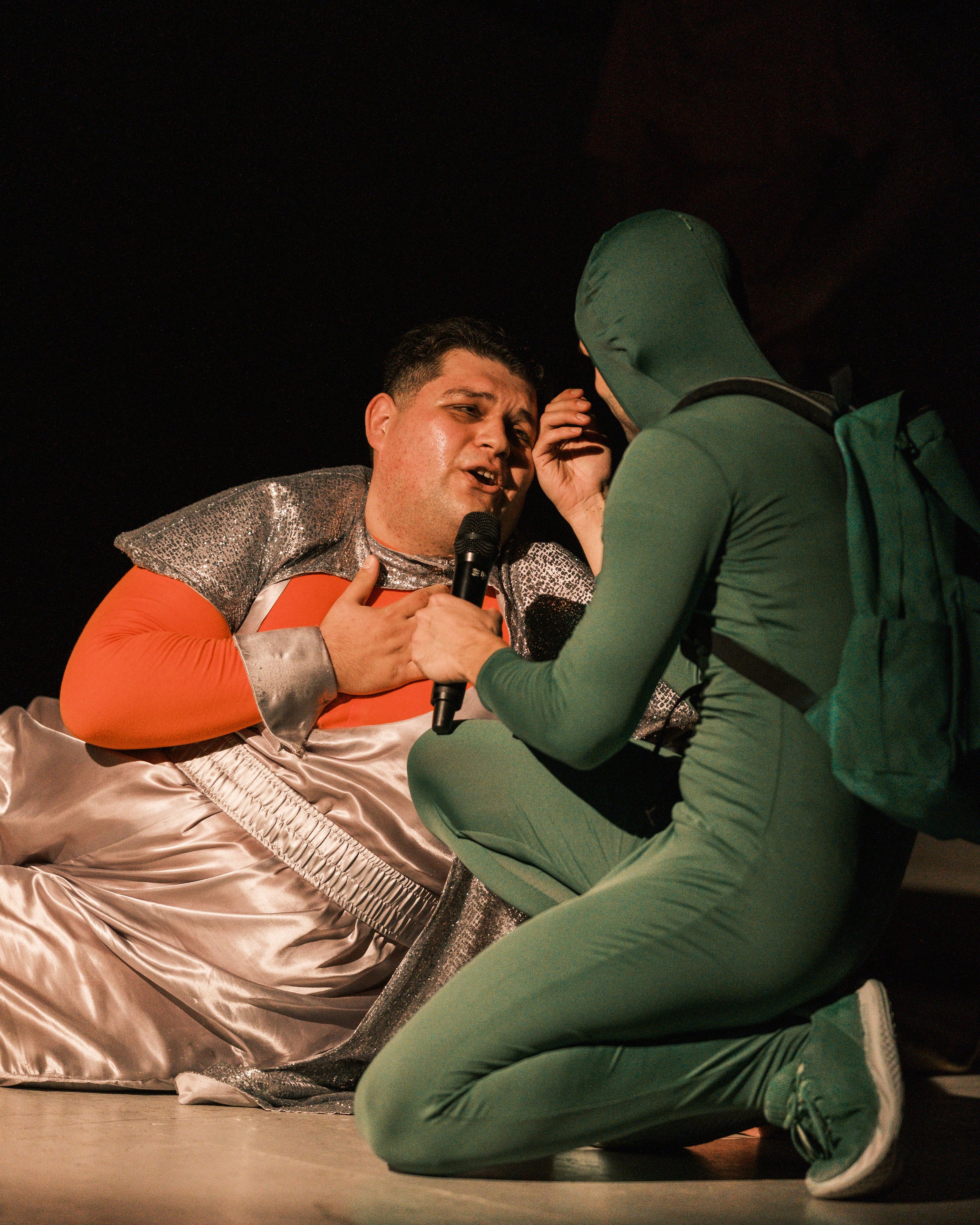 A performer in a shiny, metallic silver and orange costume with curly hair singing into a microphone while another person, dressed in a green hoodie and pants, kneels next to them.