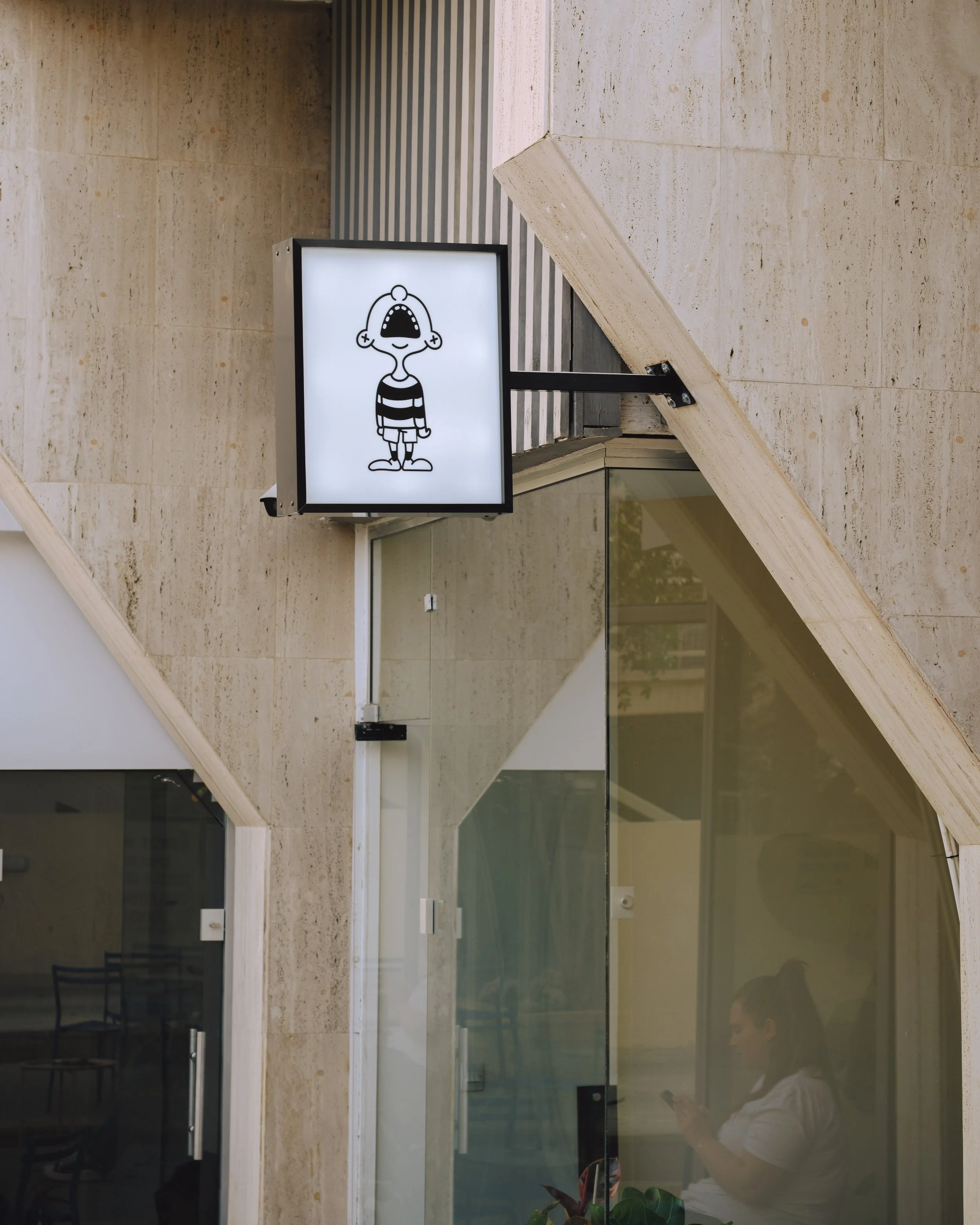 A white illuminated sign with a cartoon character of a boy with a large mouth, wearing a striped shirt, hanging from a wall outside a building. The background includes stone walls, glass windows, and reflections of an indoor person holding a phone.