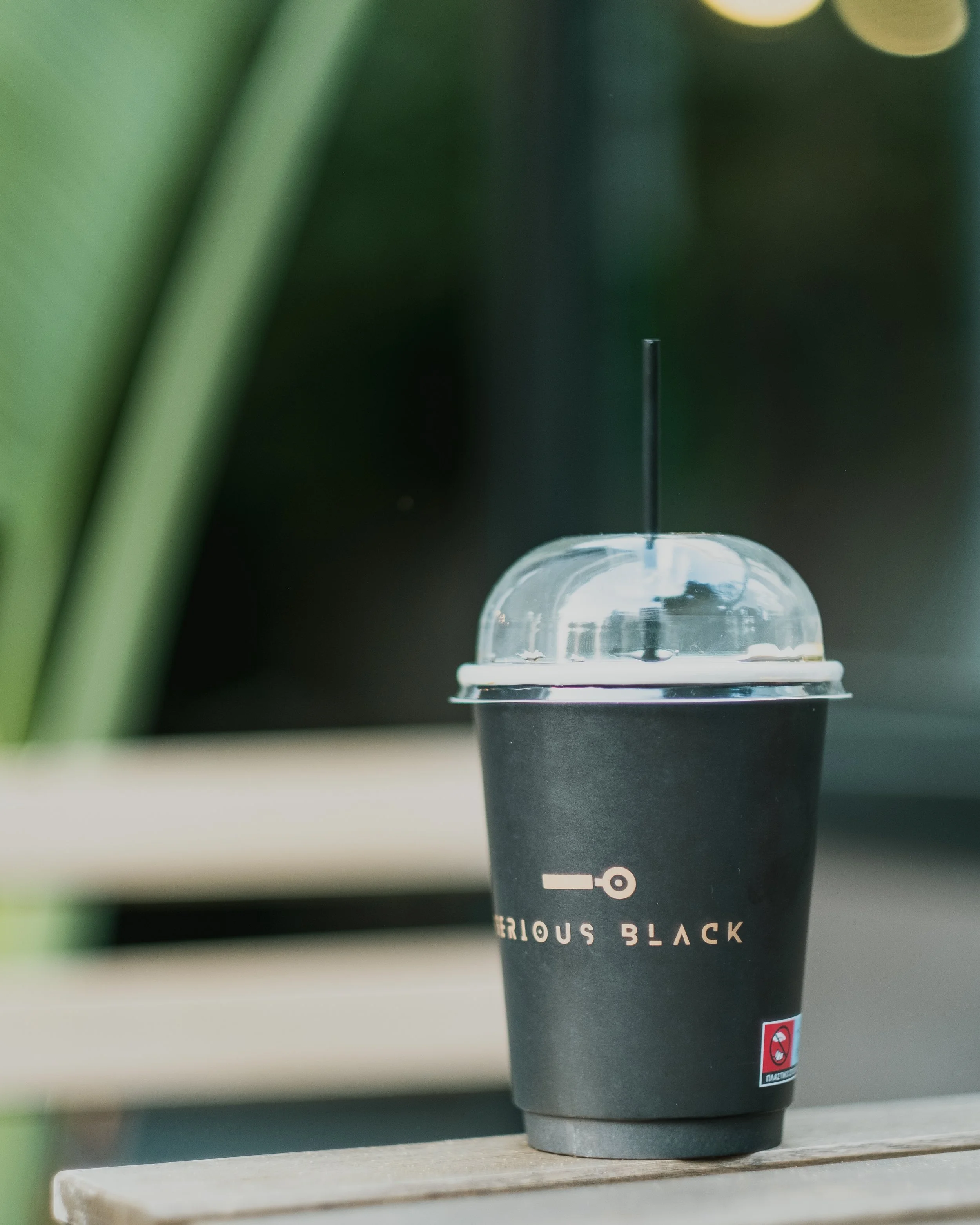 Close-up of a black disposable cup with a clear plastic lid and a black straw, placed on a wooden surface. The cup has printed text that reads 'SERIOUS BLACK' and a small warning symbol near the bottom. The background includes blurred green plant ste