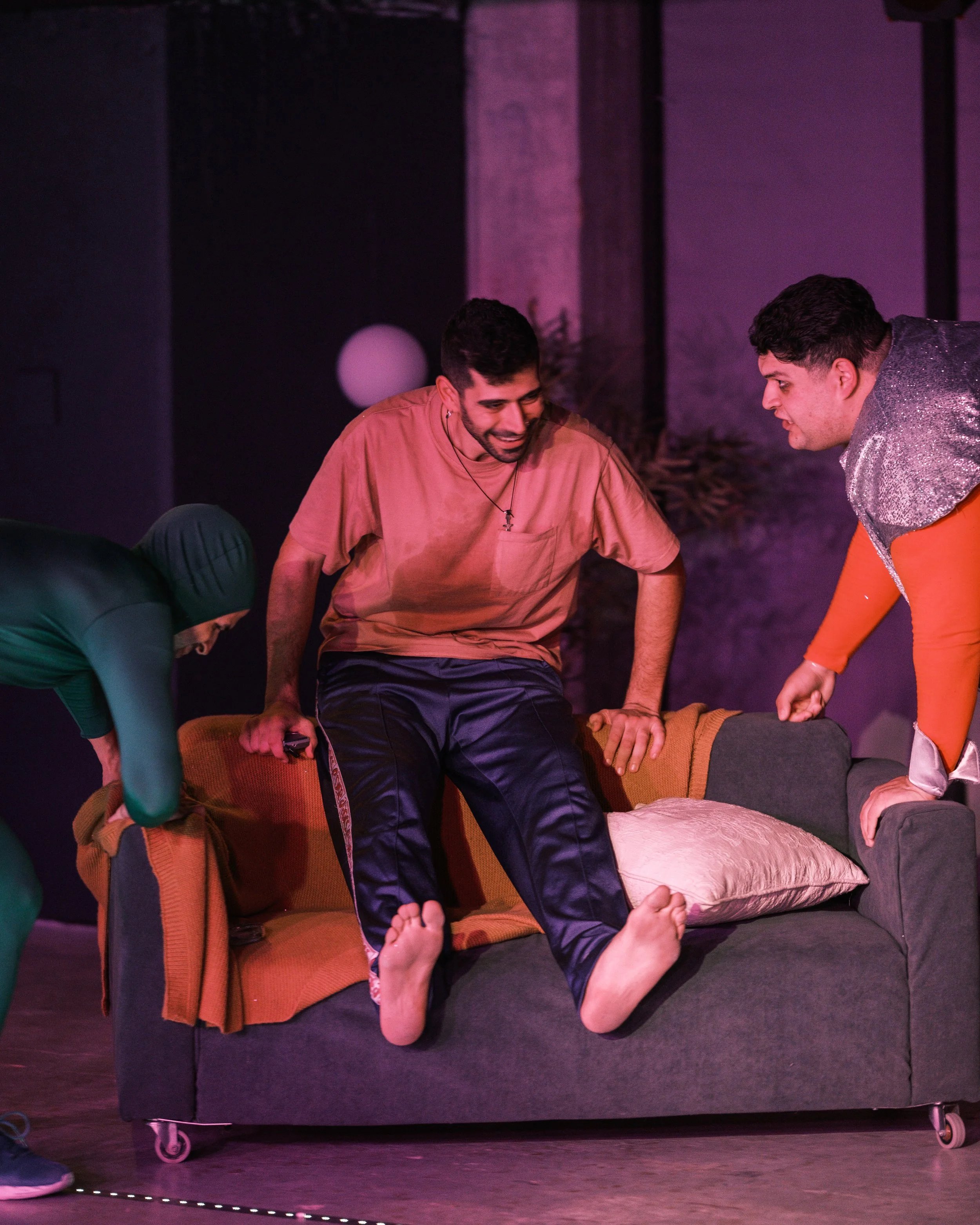 People performing a theatrical scene on stage, with a pink-lit background and props like a couch and a mannequin.