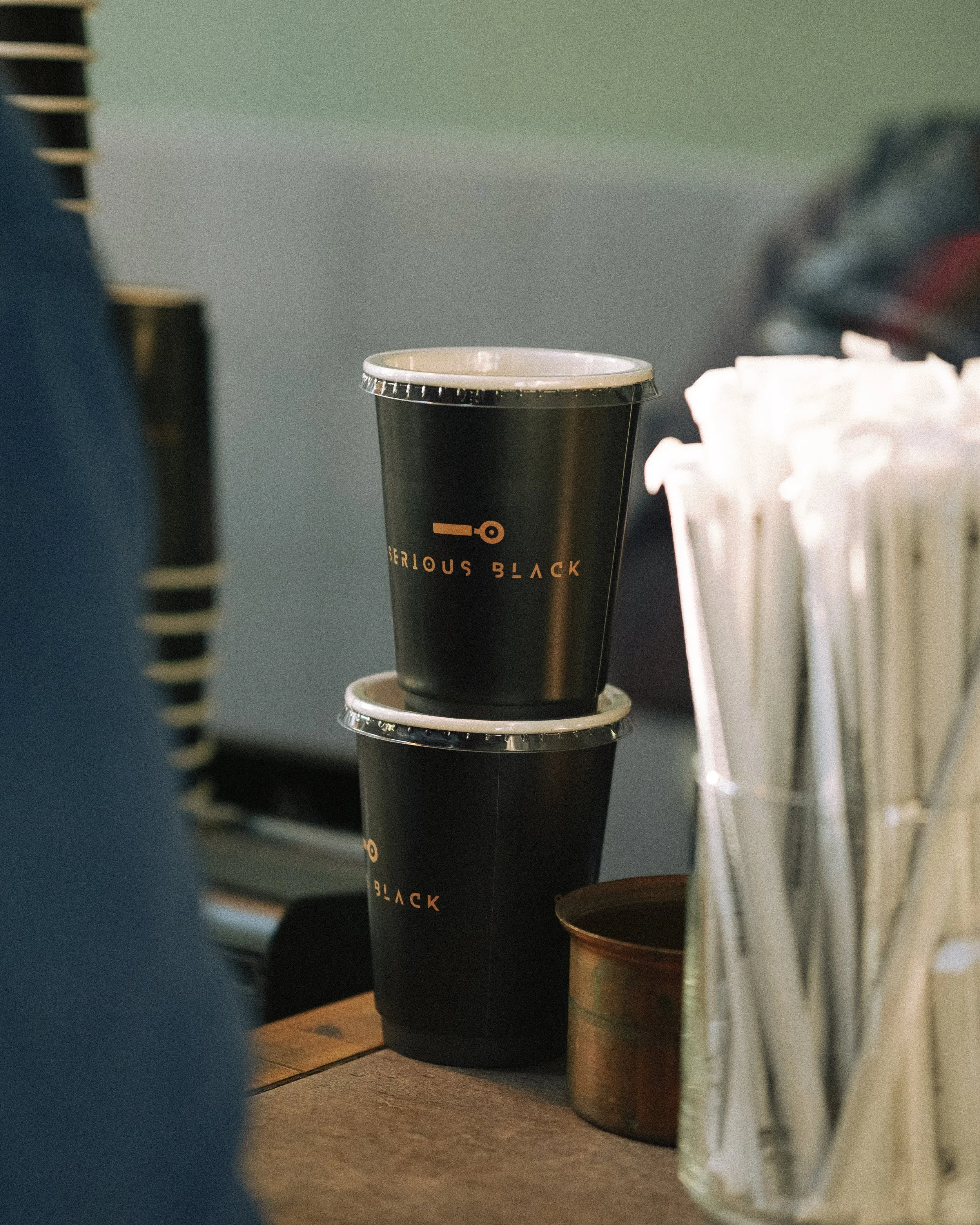 Close-up of two black disposable coffee cups stacked on top of each other on a wooden surface, with a blurred background that includes a pile of white paper napkins and other objects.