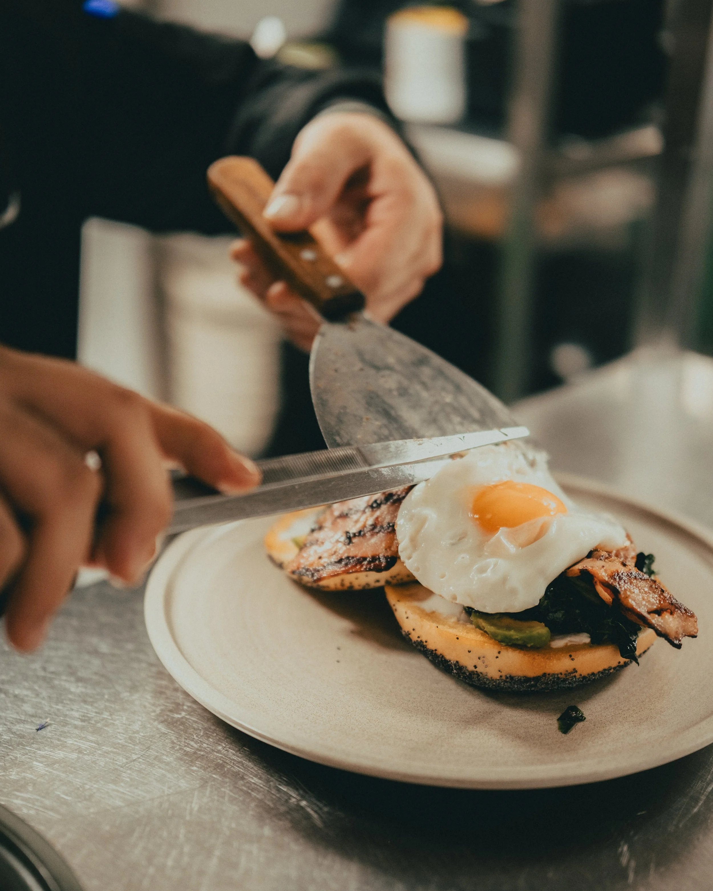 Close-up of a plate with a breakfast sandwich being cut with a knife, featuring a fried egg, grilled chicken, and vegetables on a toasted bun.