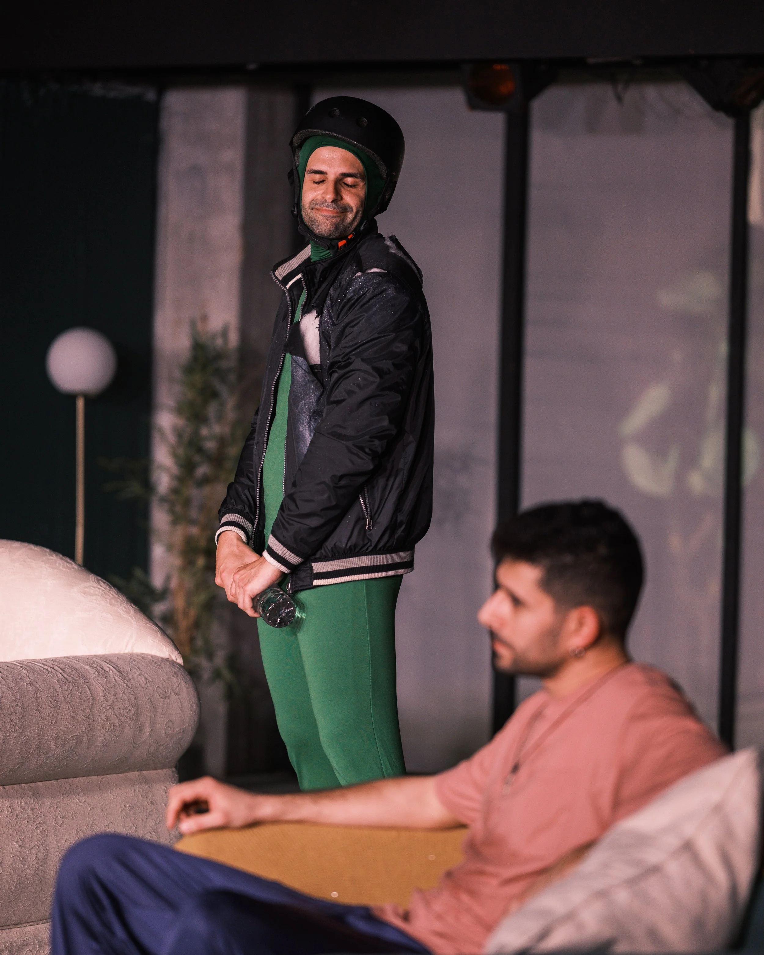 A man wearing a motorcycle helmet, jacket, and green pants standing outdoors at night, smiling with closed eyes, holding a water bottle. Another man with a beard and pink shirt is sitting nearby, with only part of his face visible.