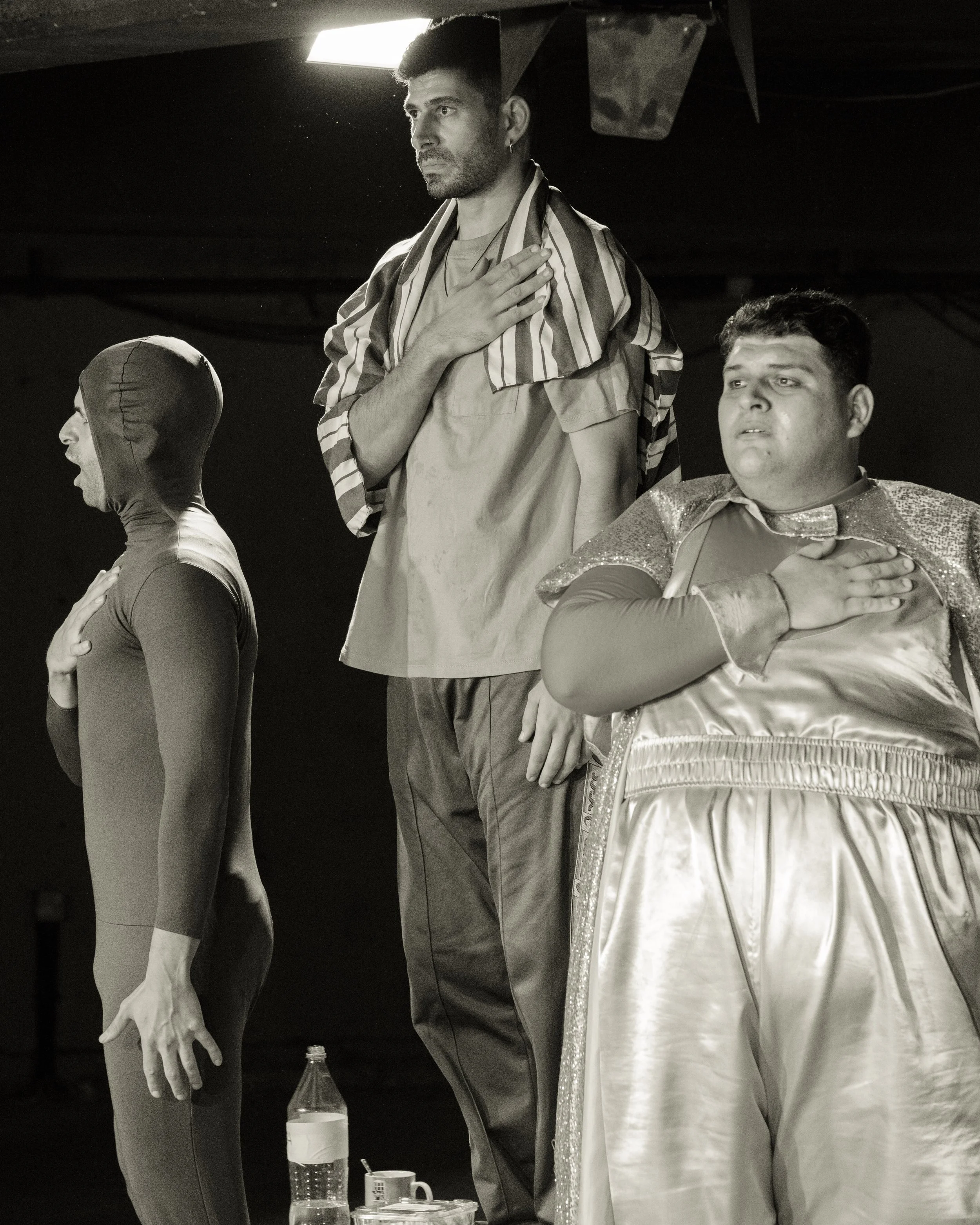Three performers standing on a stage with their right hands over their hearts, one dressed as a mime, one wearing a shiny costume, and one with a striped fabric over his shoulders. The scene is in black and white.
