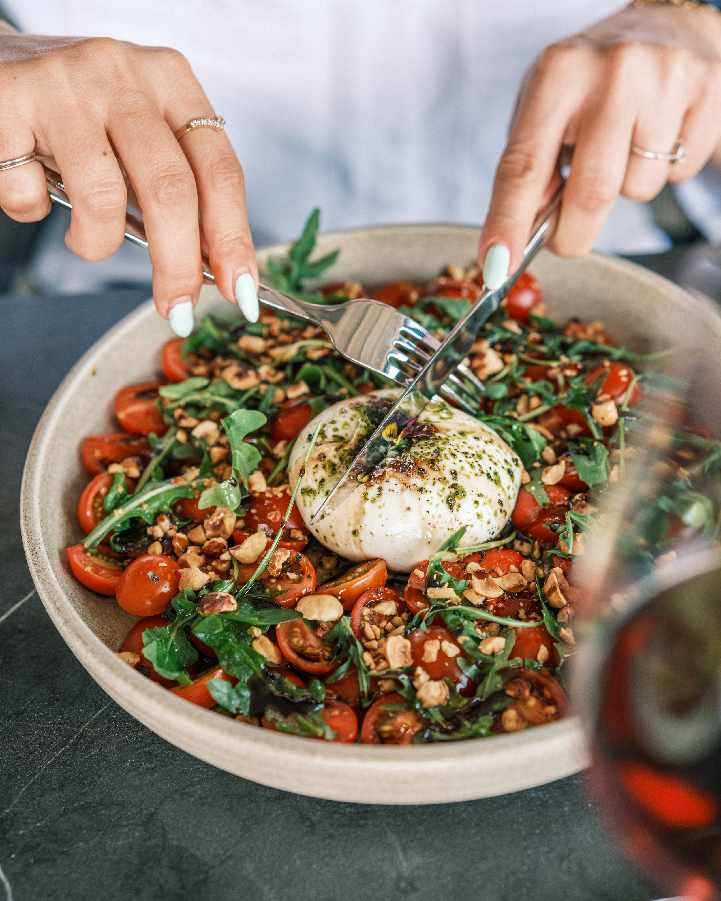 A person making a salad with cherry tomatoes, arugula, chopped nuts, and a poached egg topped with green sauce, using a fork and spoon.