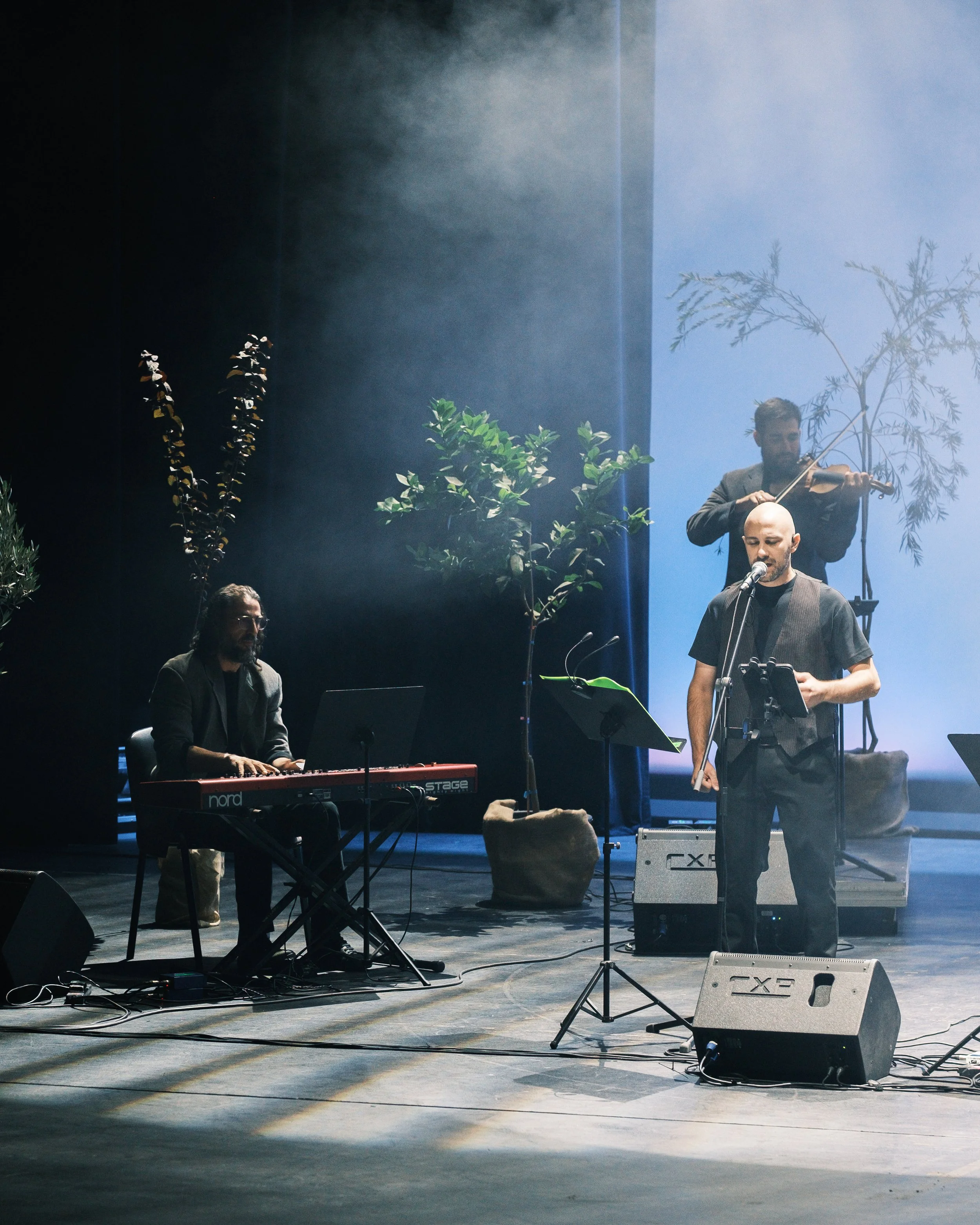 Musicians performing on stage with a keyboard, a singer, and a violin player surrounded by stage plants and equipment, with atmospheric lighting.