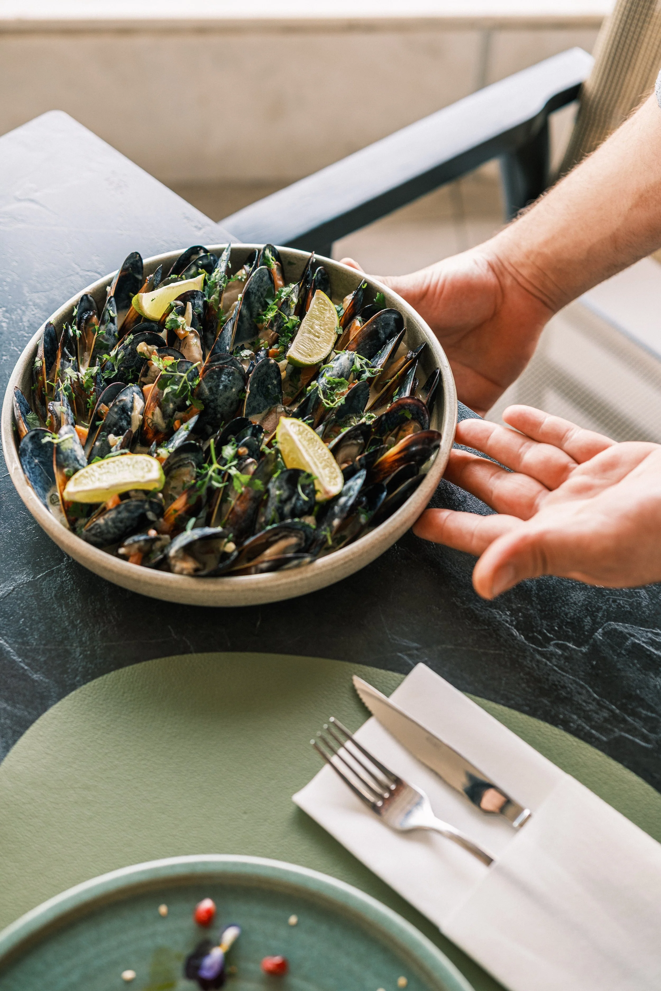 A person serving a bowl of cooked mussels garnished with herbs and lime wedges on a table with cutlery and plates.