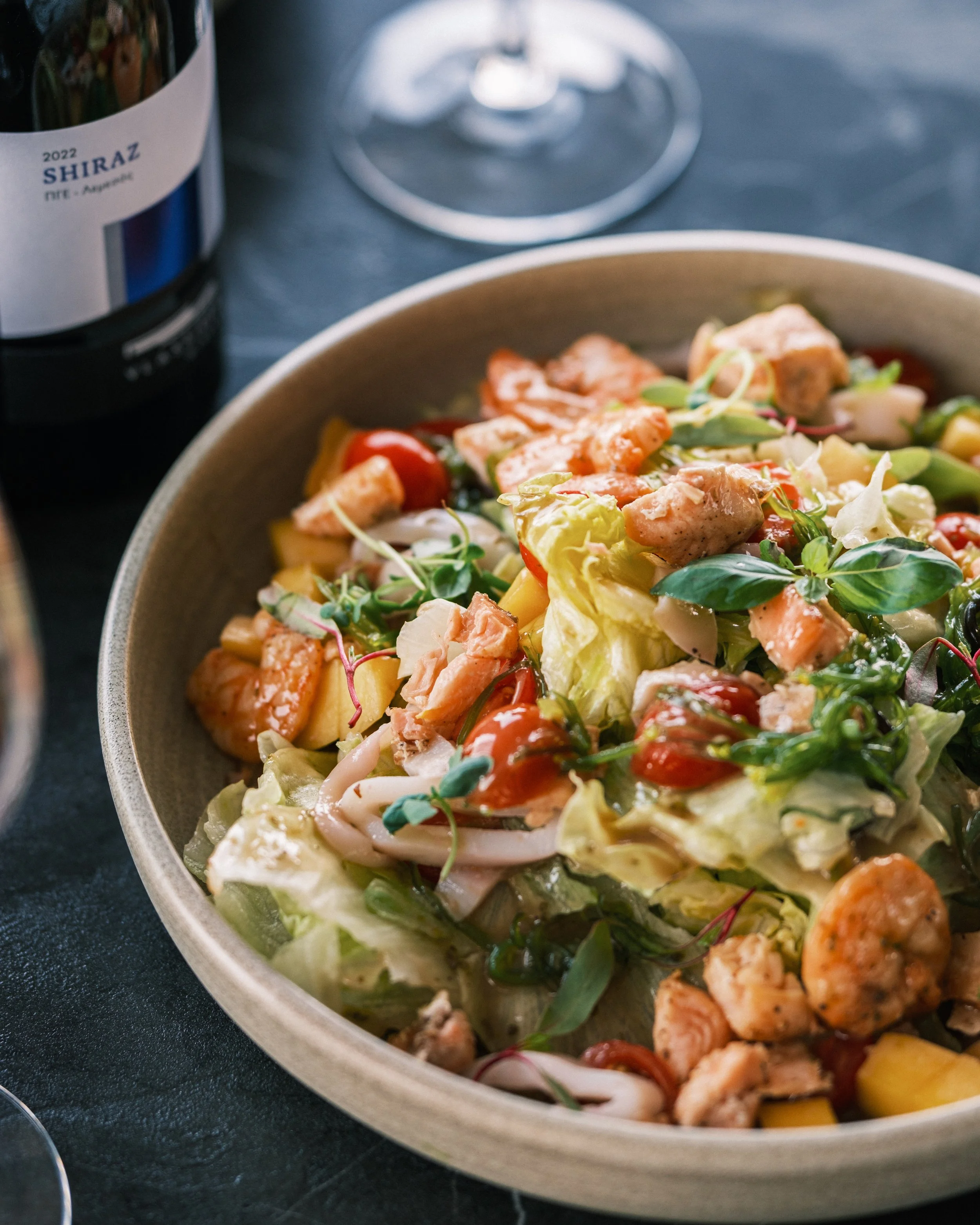 A bowl of mixed salad with lettuce, cherry tomatoes, diced chicken, and sliced vegetables on a dark surface, with a glass of wine and a bottle of Shiraz wine in the background.