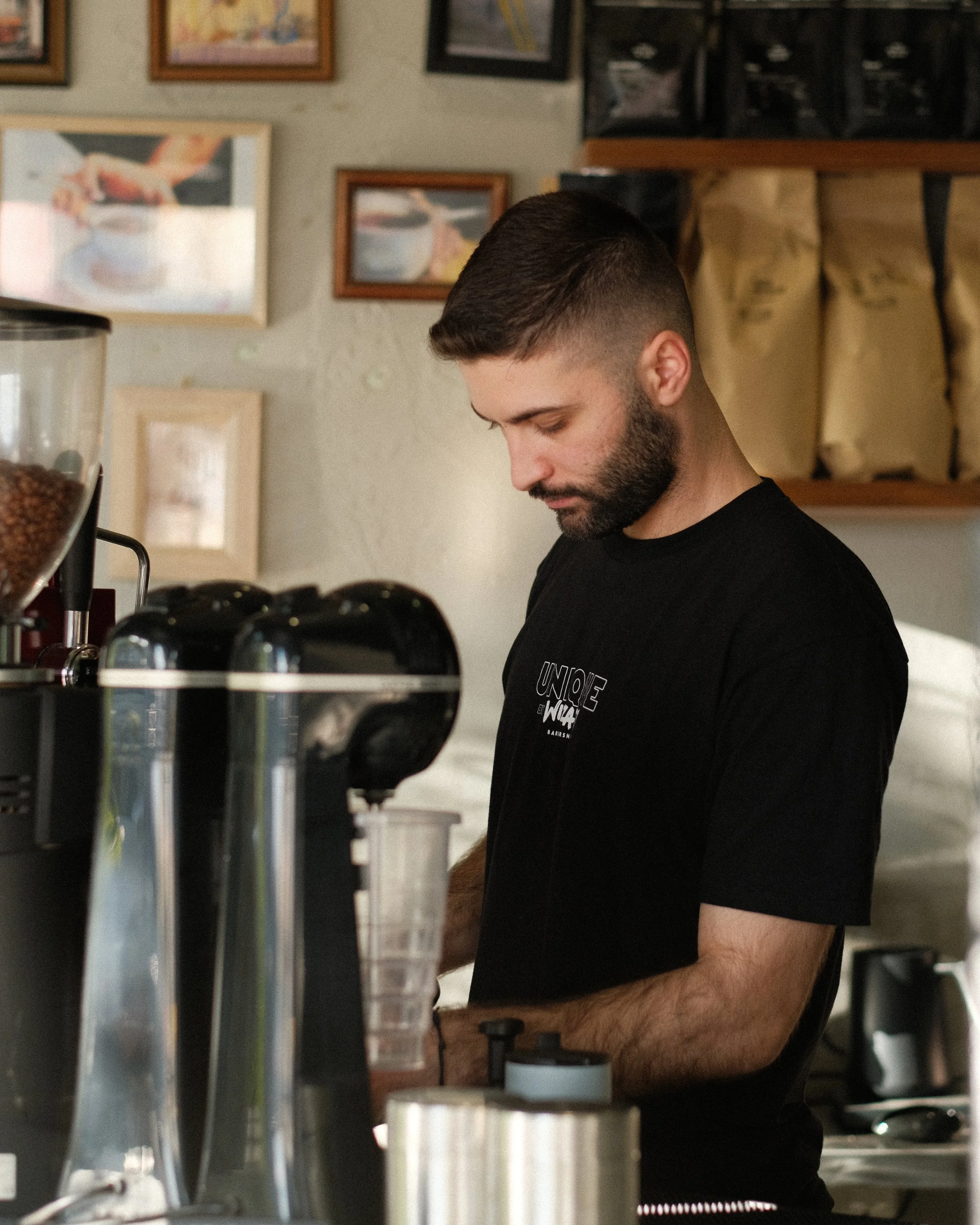 A man with short dark hair and a beard standing in a coffee shop. He is wearing a black T-shirt with white text and is focused on his work. Behind him are framed photographs on the wall and coffee bags on shelves.