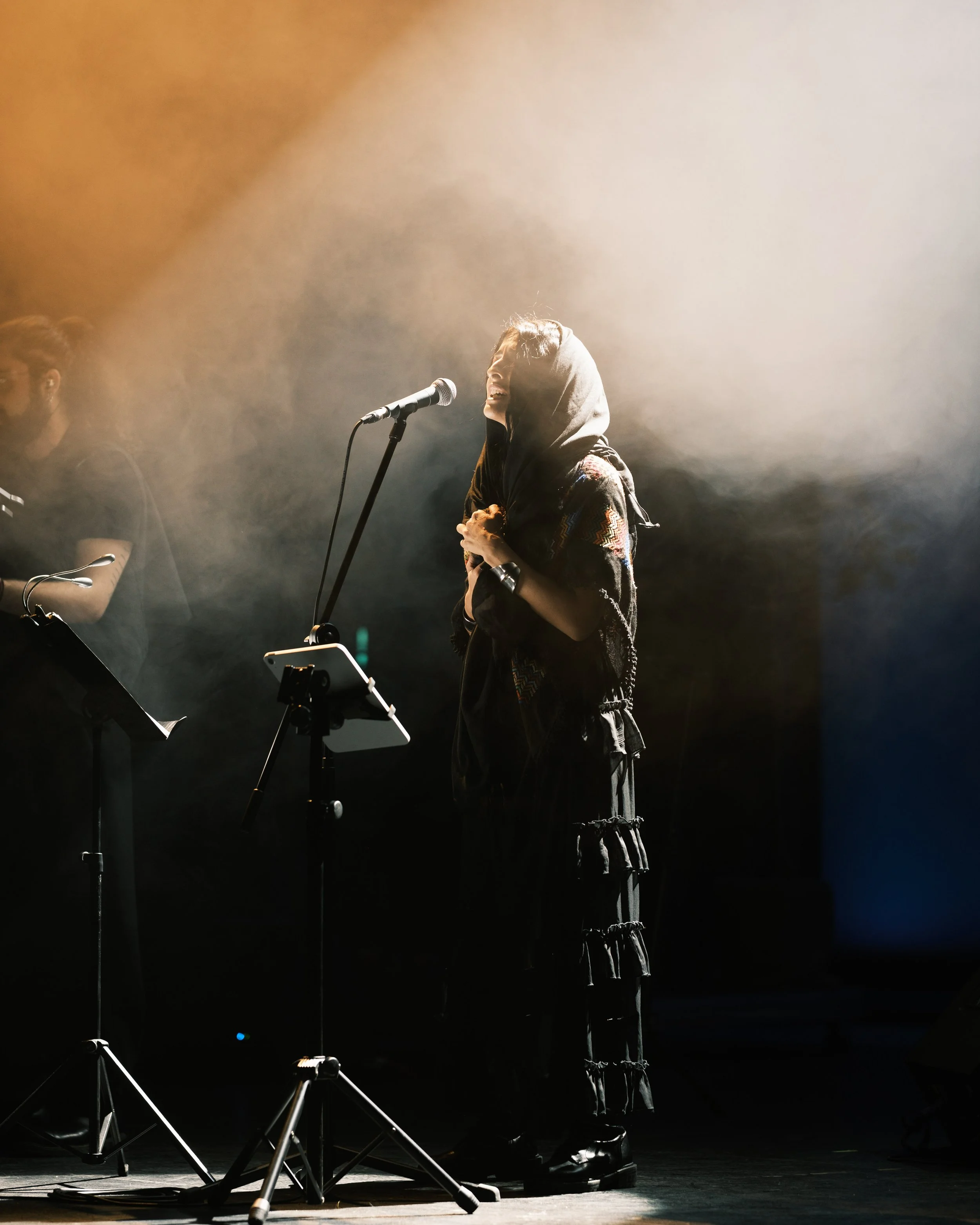 A woman wearing a hoodie, smiling, and singing into a microphone on stage with stage lights creating a hazy atmosphere.