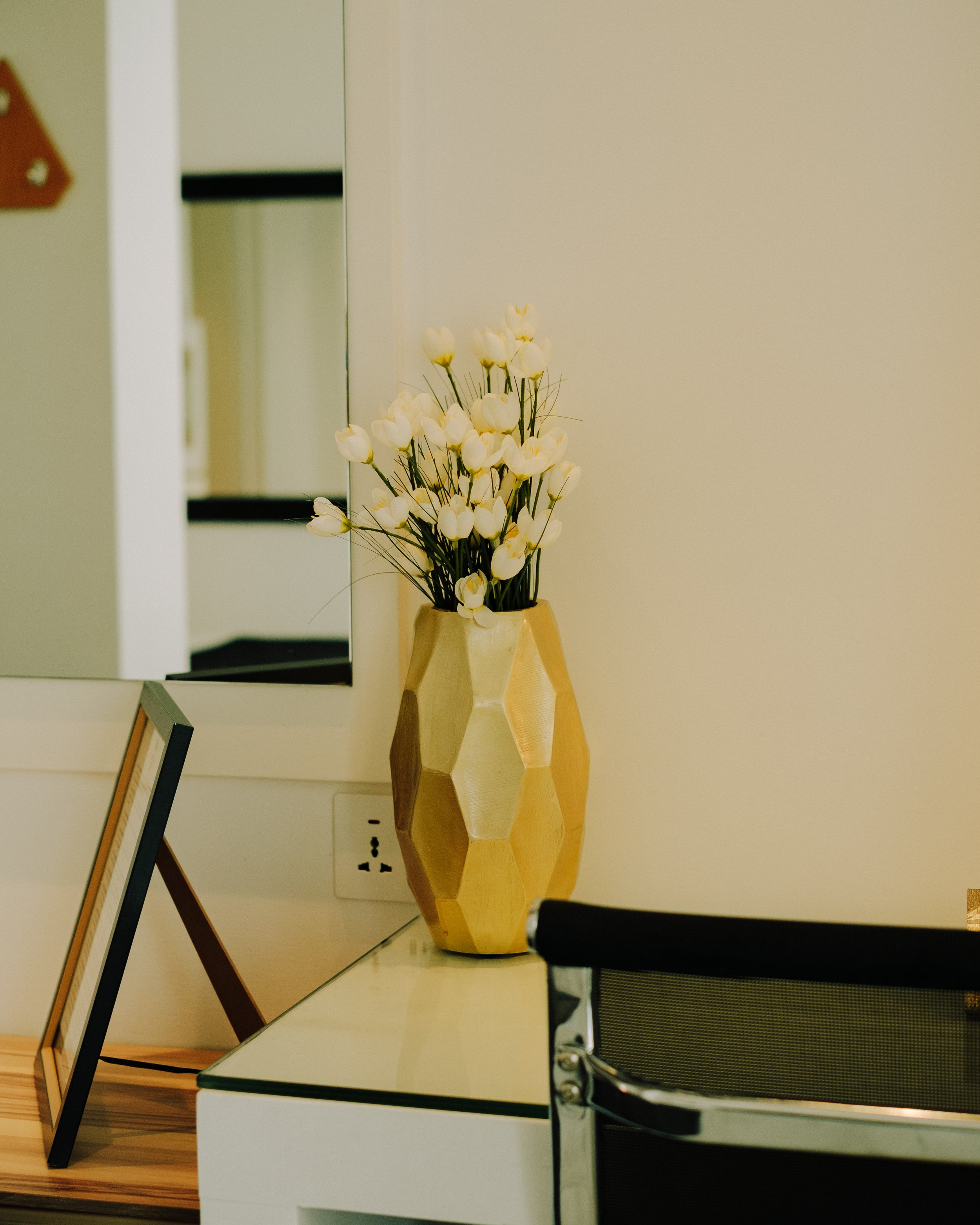 A decorative beige geometric vase with white tulips on a table in a modern office or room setting, with a mirror behind it reflecting part of the room and a power outlet on the wall.