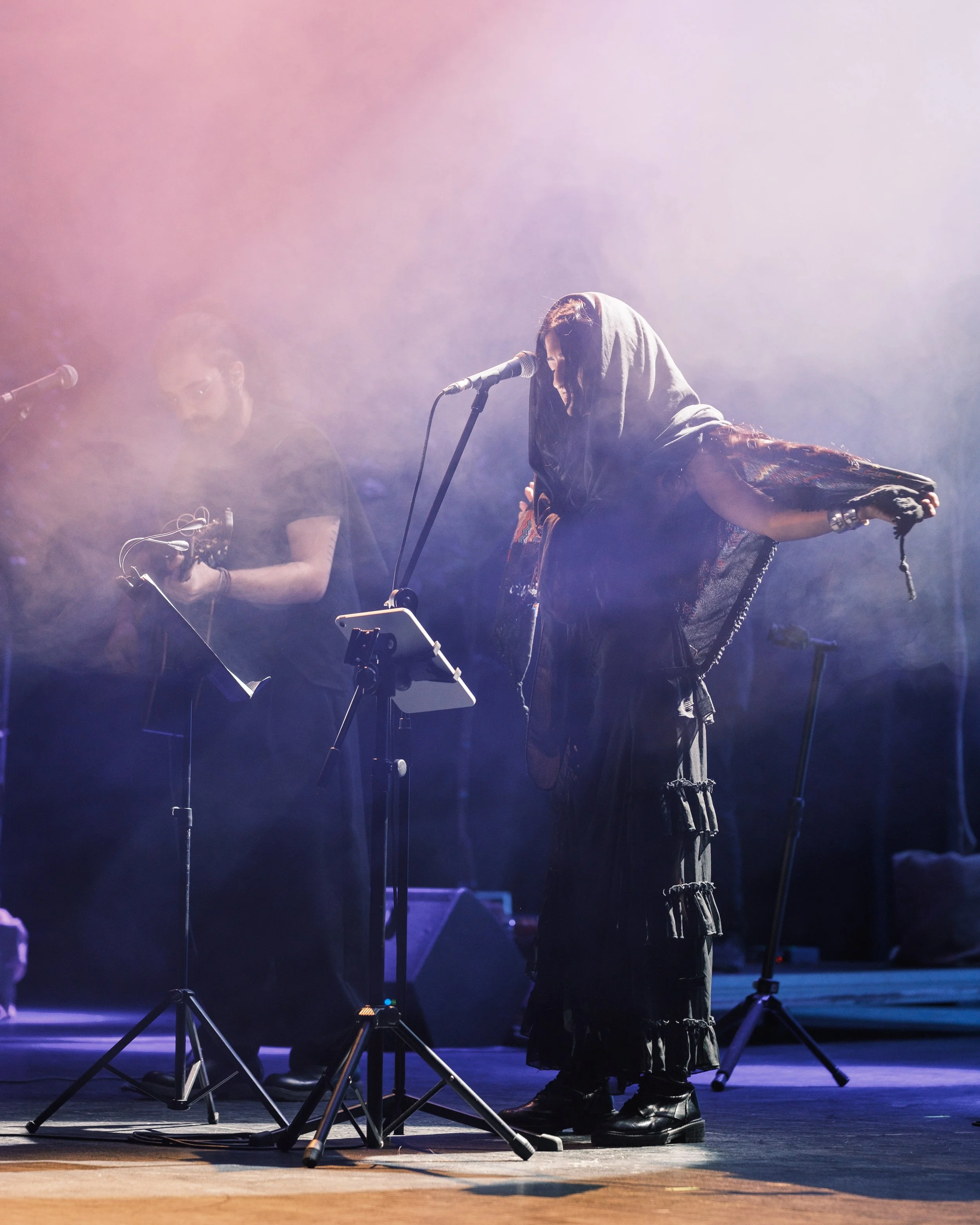 A woman with long dark hair wearing a hooded shawl and a dark dress, standing on stage at a microphone with her head bowed and her arm extended, holding a chain. Two men with equipment are in the background, on stage, in a foggy, colorful lighting se
