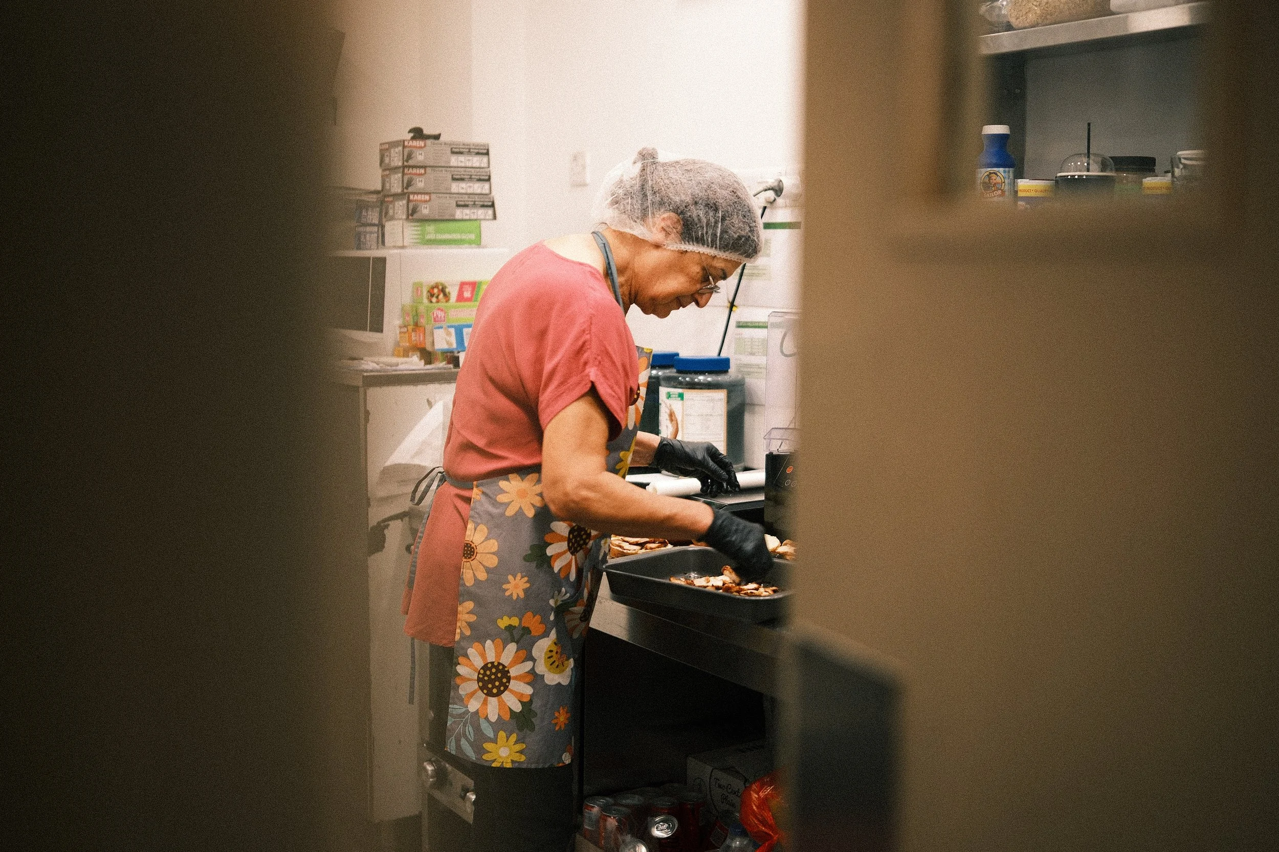 A woman with a shower cap, glasses, and a floral apron prepares food in a kitchen, seen through a small opening.