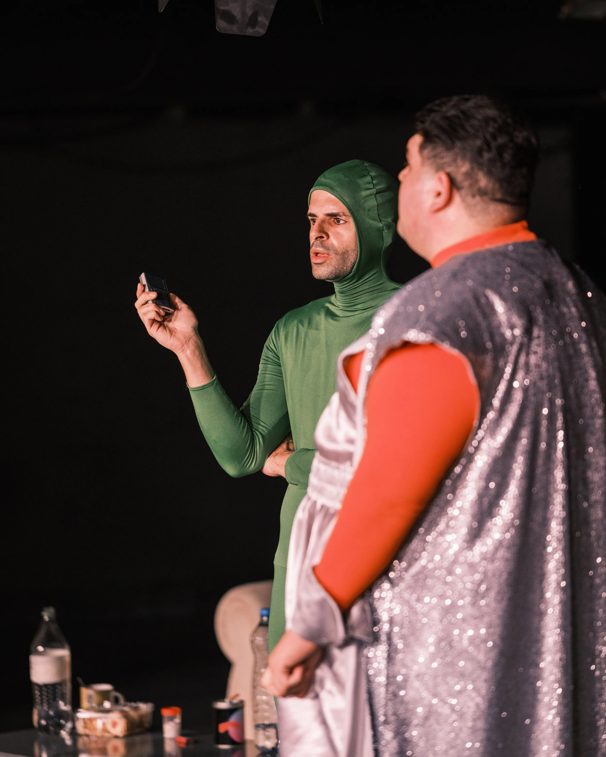 Two men standing next to each other, one wearing a green bodysuit with a hood and the other wearing a shiny, sparkly silver jacket with orange accents. The man in the bodysuit is holding a small device, possibly a phone or remote, and appears to be f