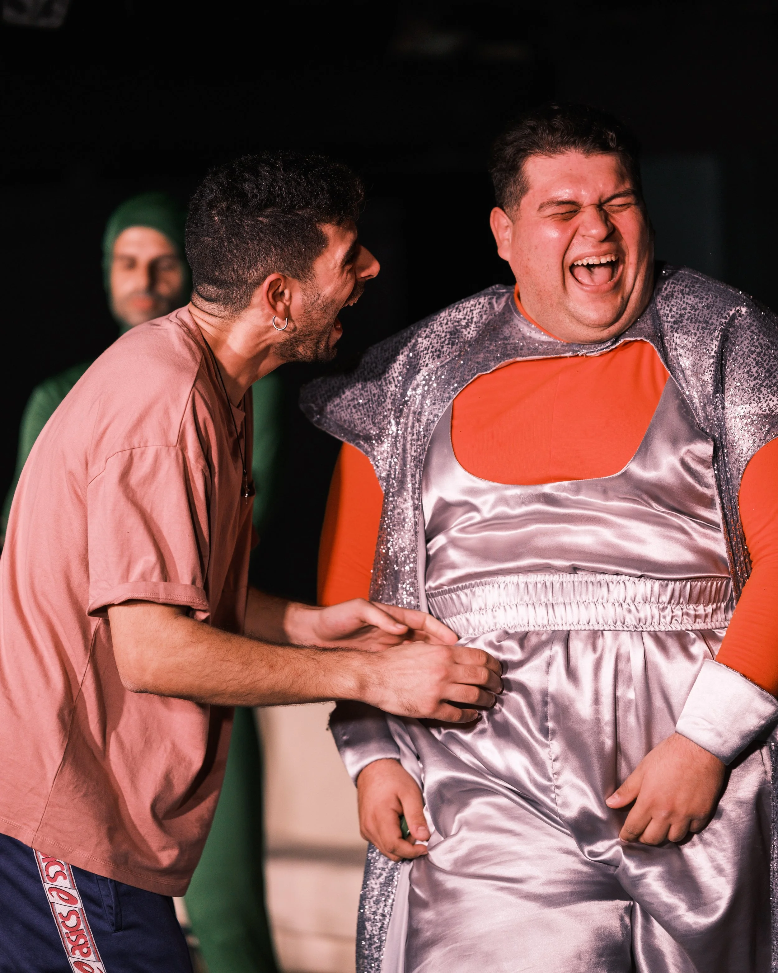 Three men laughing and smiling. The man on the right is wearing a shiny silver and orange outfit, and the man in the middle is wearing a pink shirt. The man on the left is partially visible, wearing a green hat.