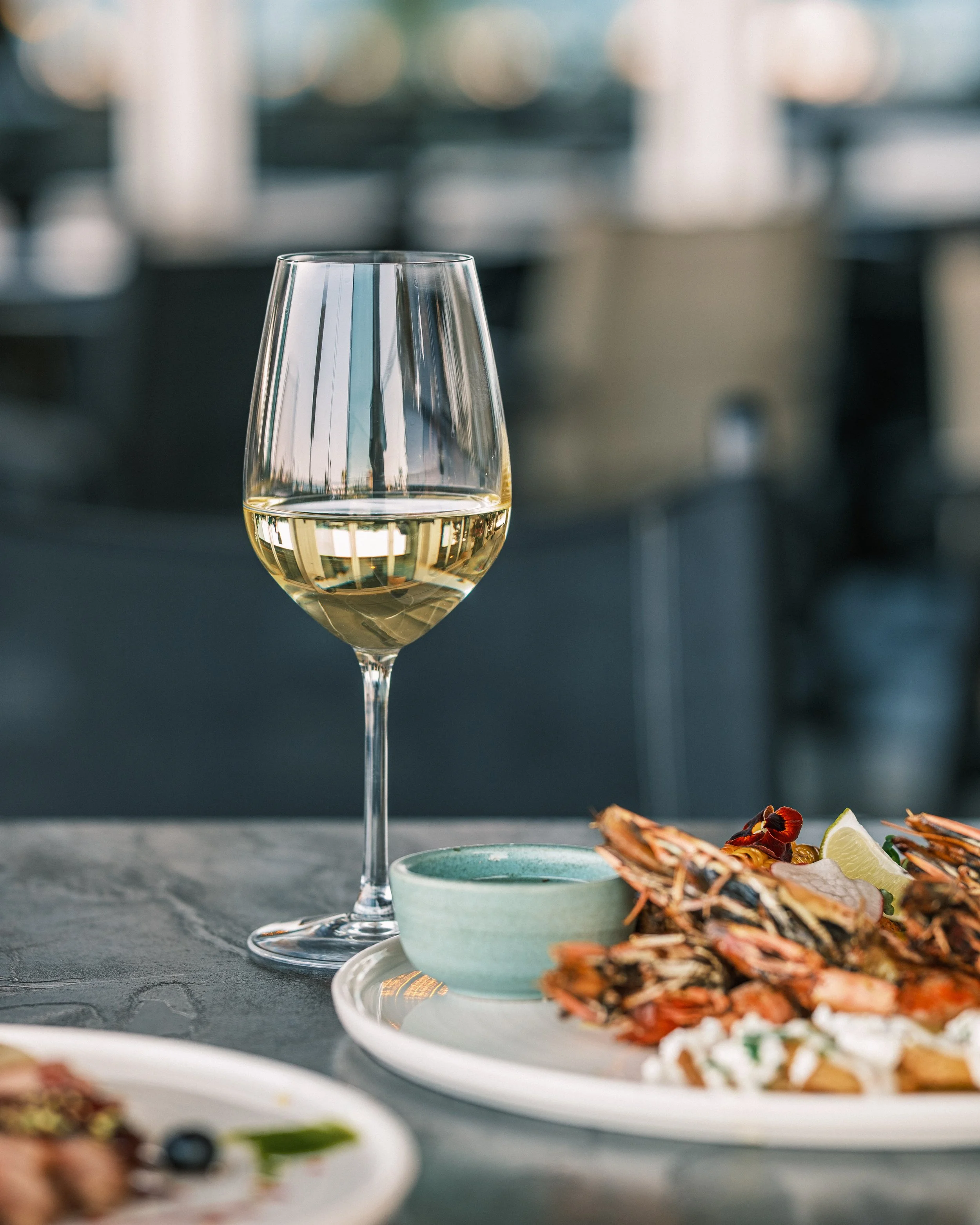 A glass of white wine on a dark surface with a blurred background. Part of a seafood platter with lobster and lemon slices is visible in the foreground.