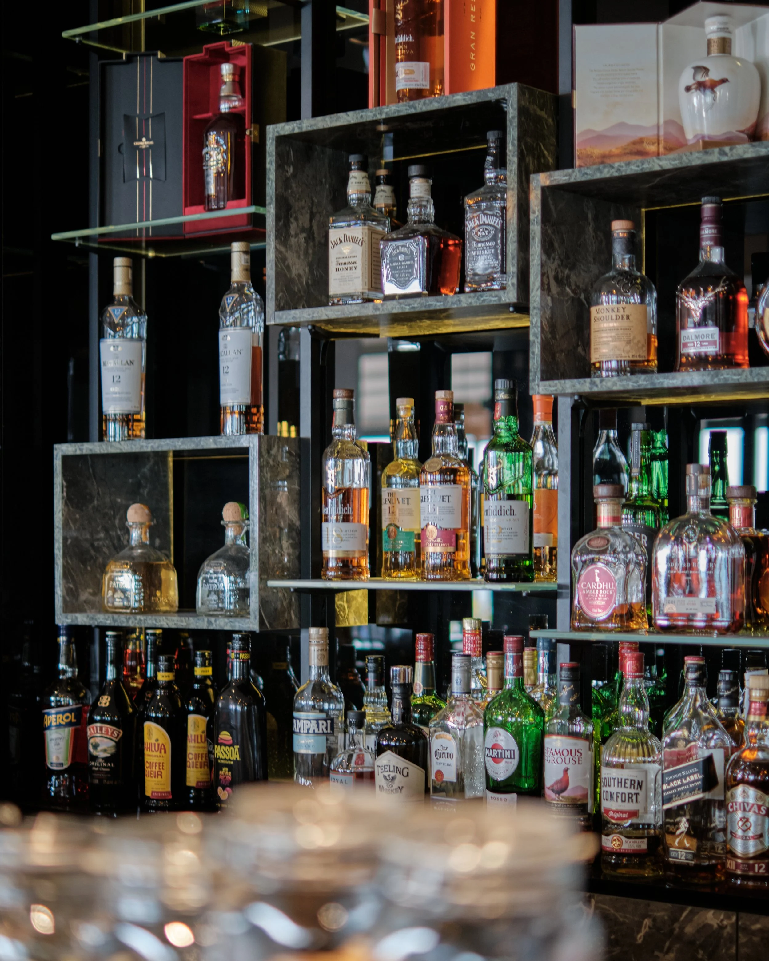 A variety of whiskey bottles on display on black metal shelves with some in cardboard boxes, next to other bottles of liquor, in a store or bar setting.