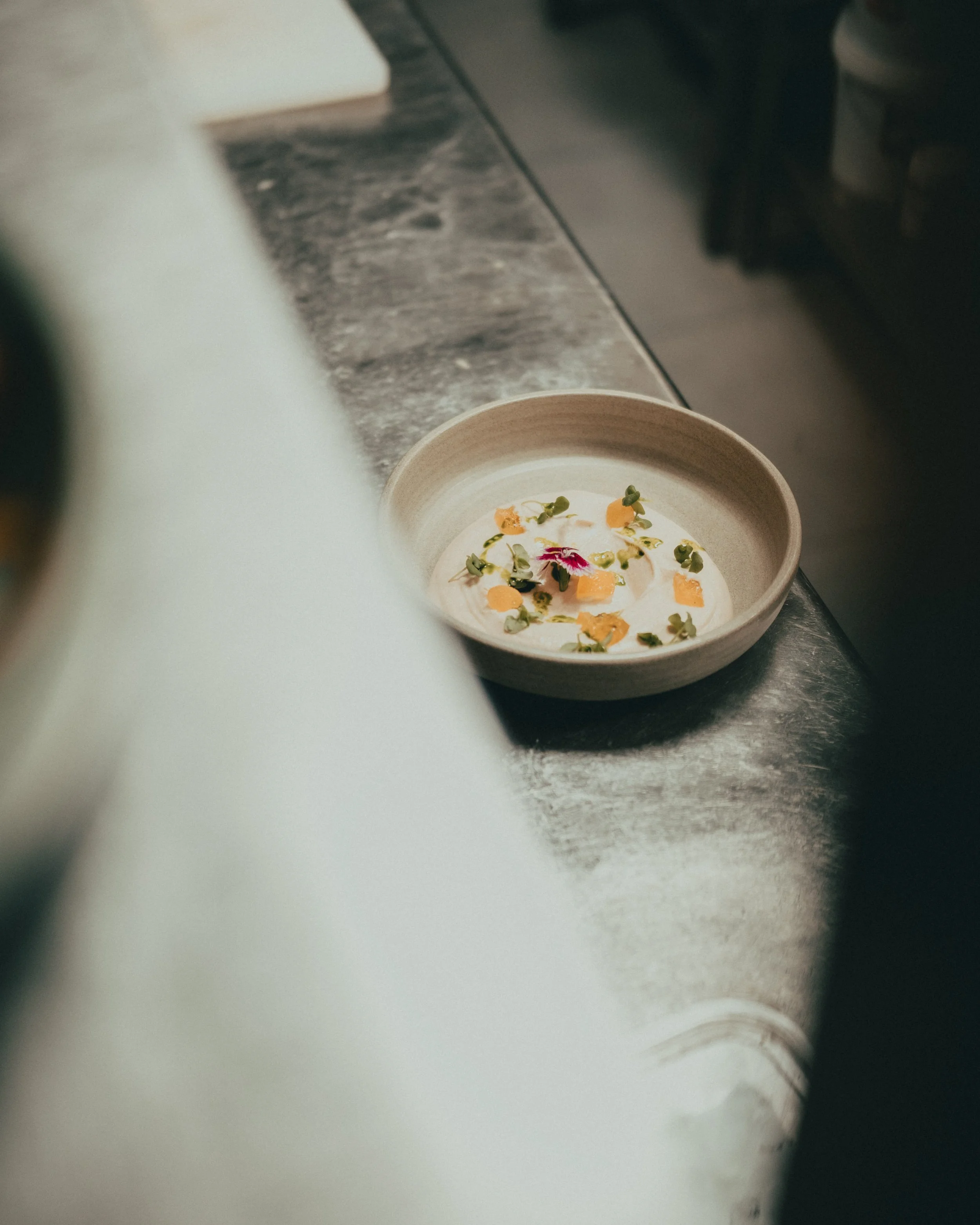 A bowl of creamy soup garnished with microgreens, orange flower petals, and a drizzle of oil, placed on a metallic countertop in a kitchen.